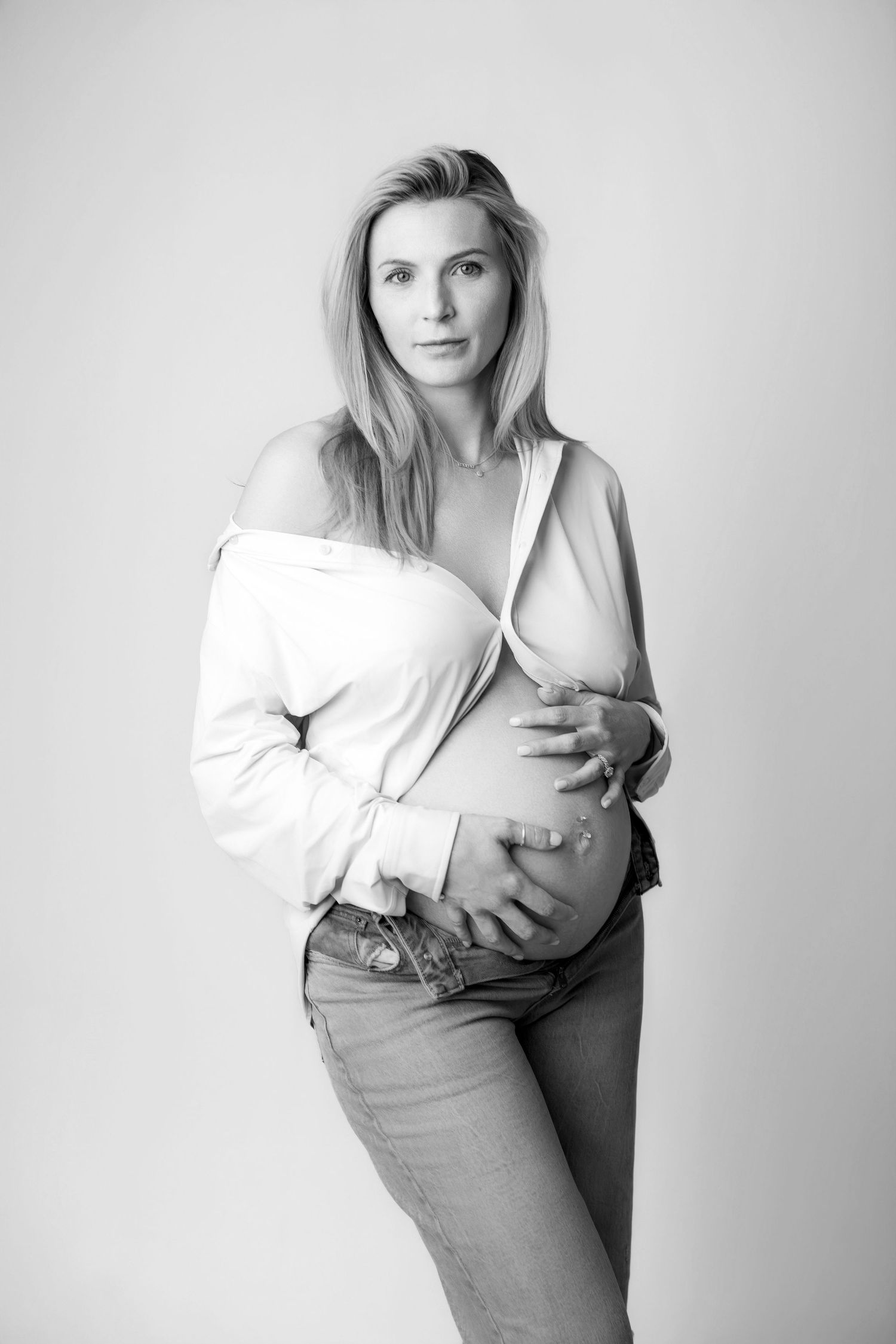 Black and white portrait of pregnant woman in open white shirt cradling baby bump.