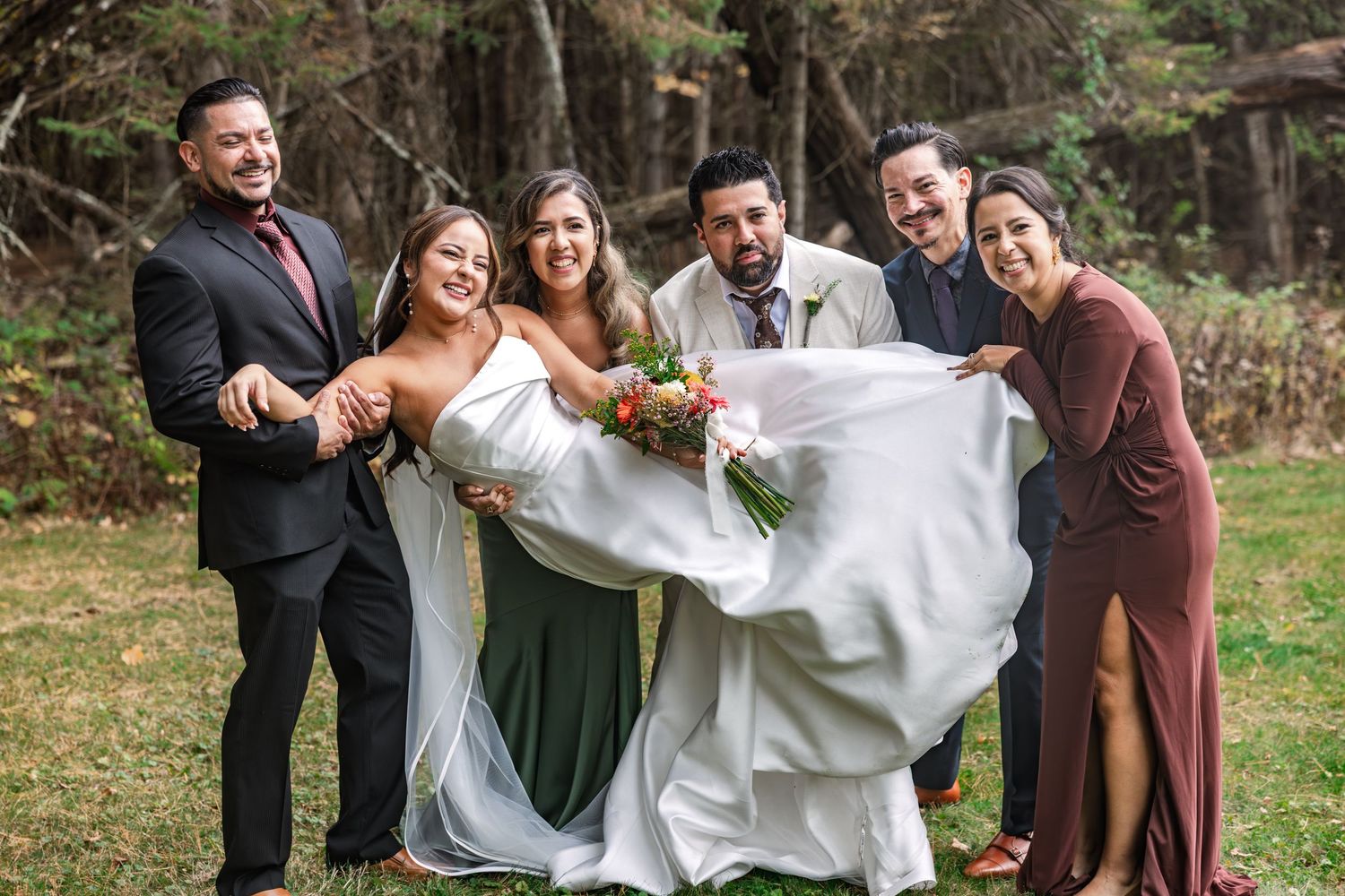 Bride and groom celebrate joyfully with wedding party in an outdoor autumn forest setting.