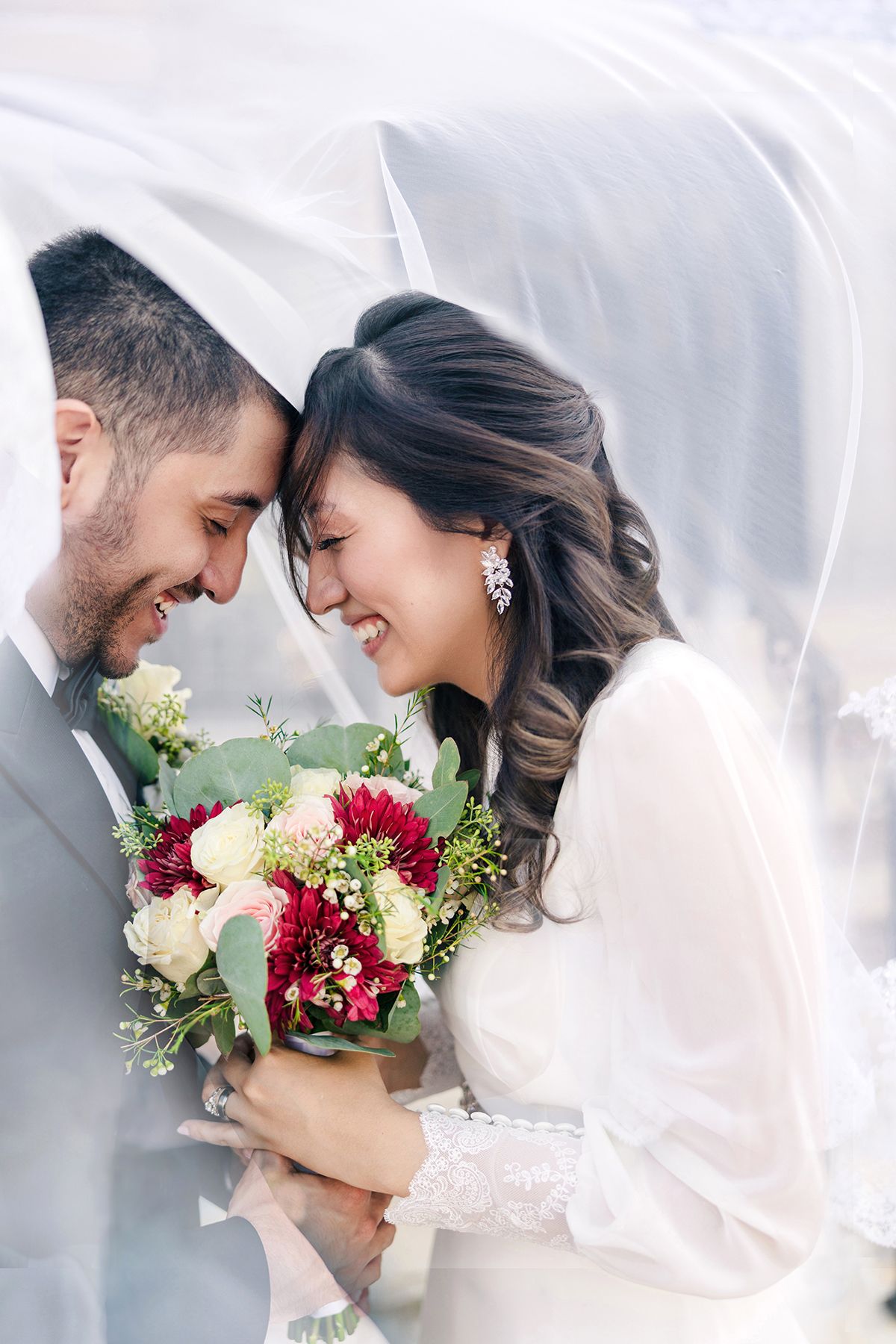 Happy bride and groom laughing together under white veil holding red and white bouquet.