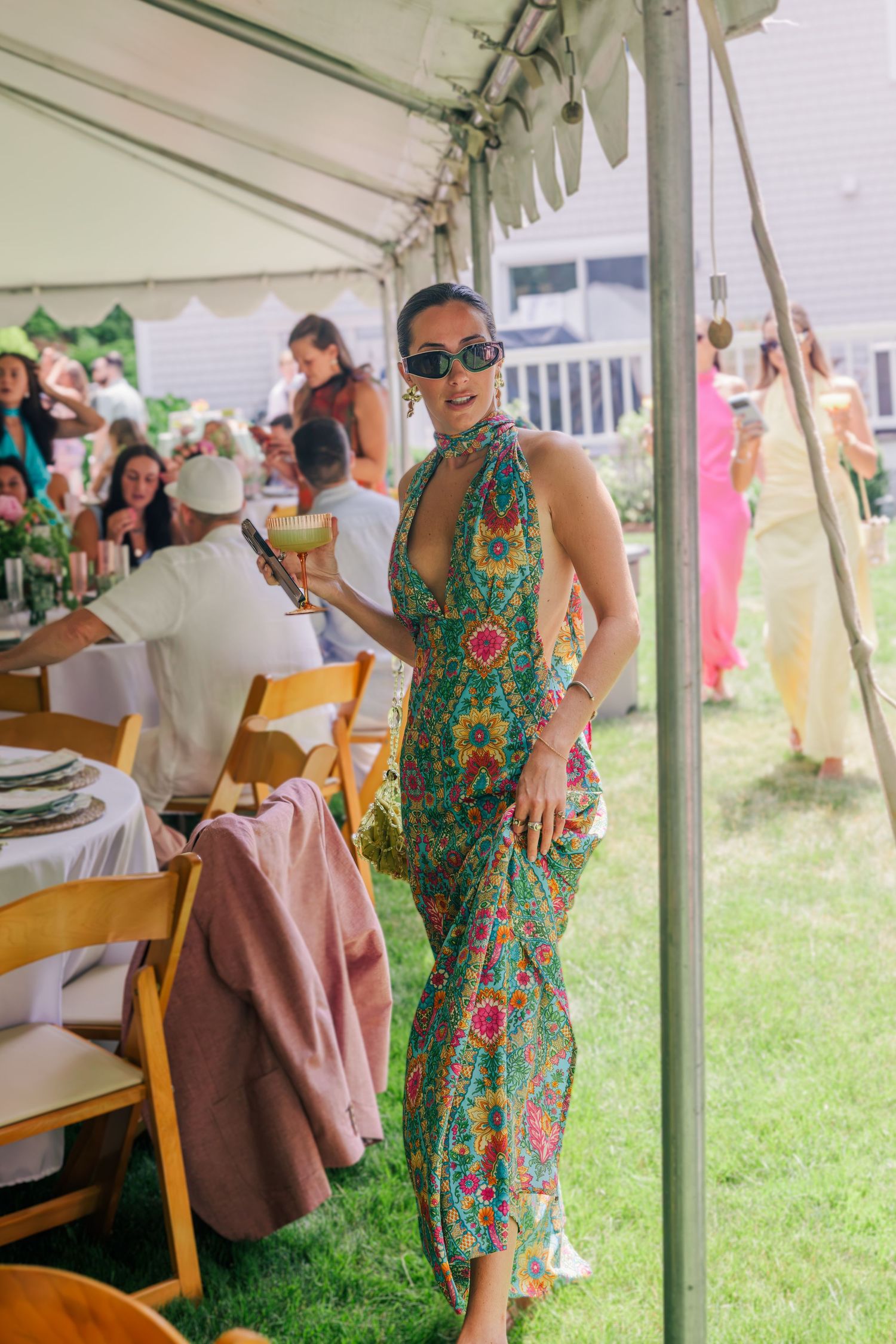 Woman in colorful floral halter dress holding cocktail at outdoor garden party under white tent.