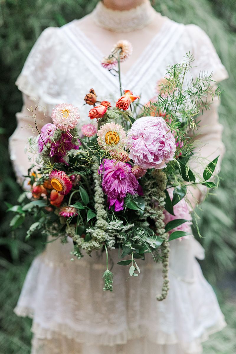 Bride holding a lush wildflower bouquet with pink peonies, dahlias, and greenery in a garden setting.