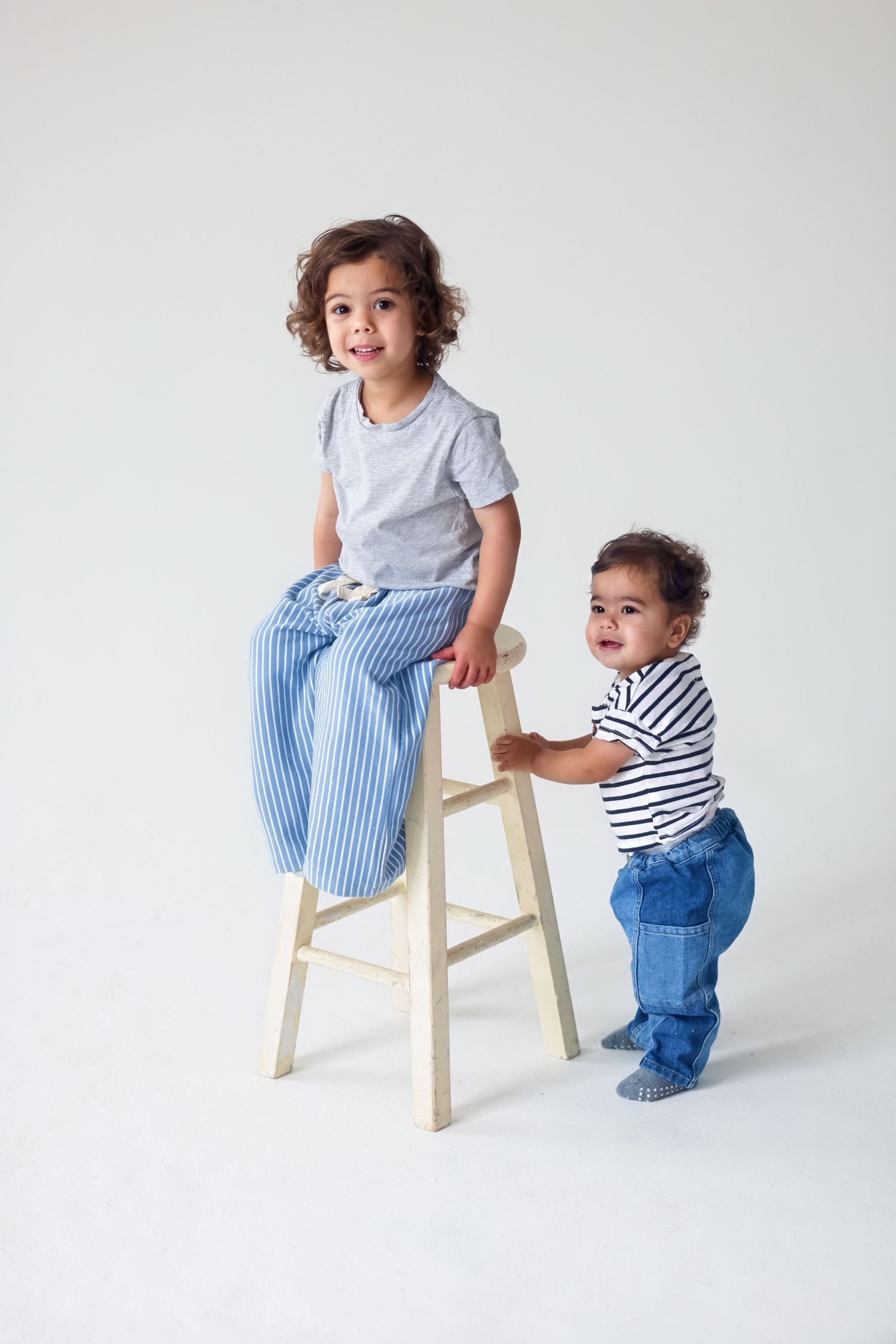 Two cute young siblings pose in a white studio, one sitting on a wooden stool and one standing.