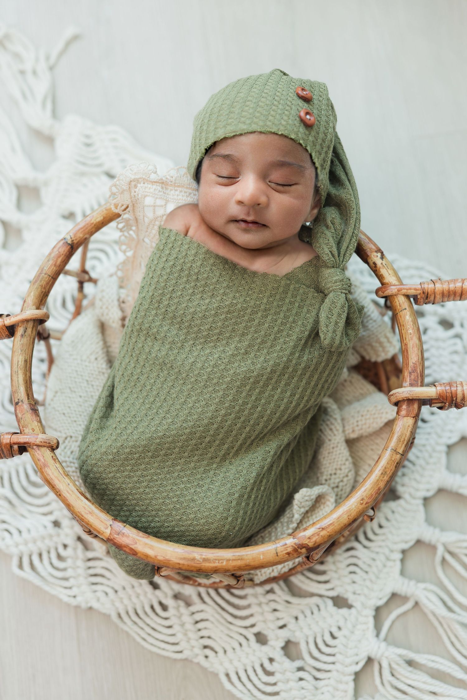 Newborn baby wrapped in green swaddle with matching knit hat sleeping in round wicker basket.