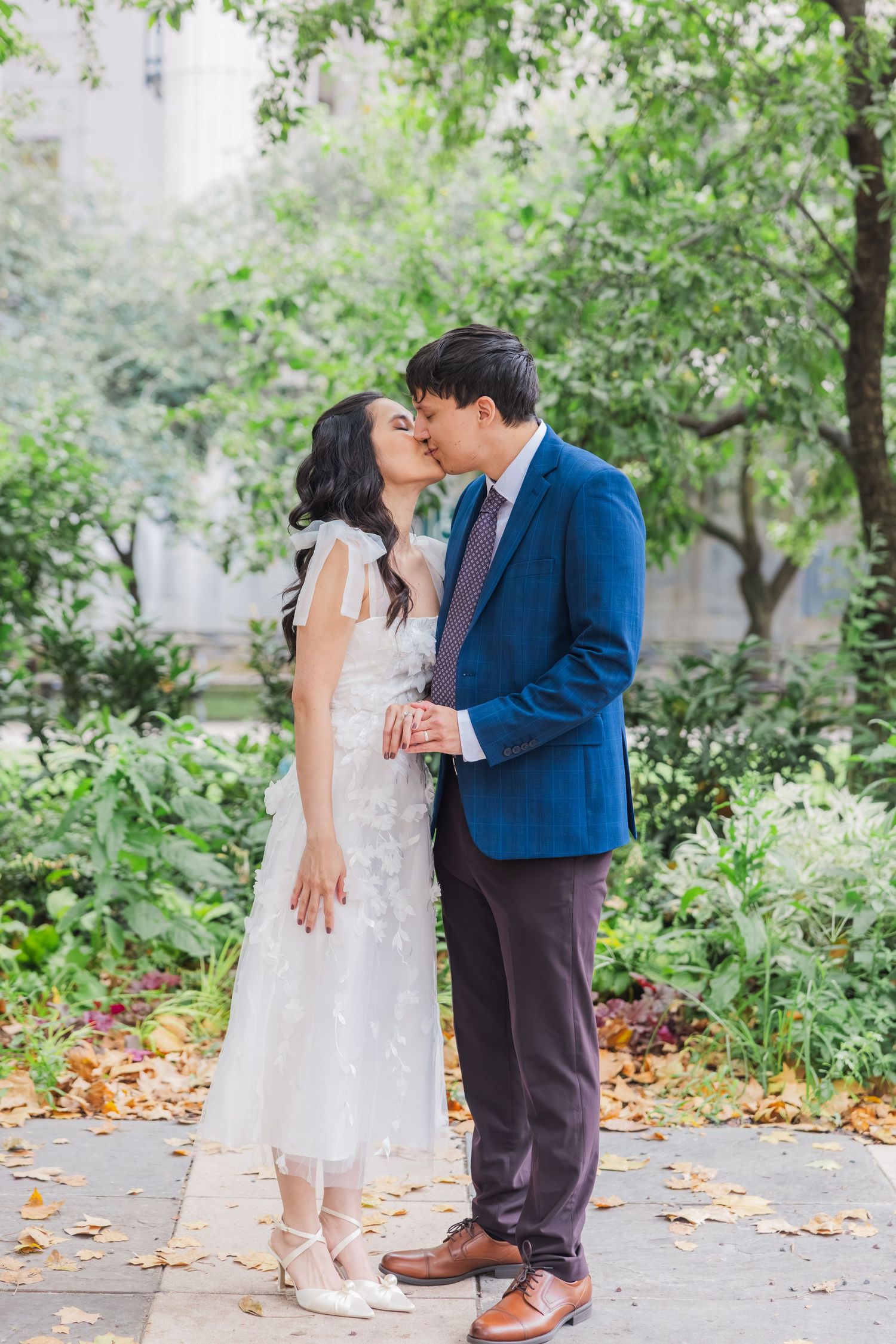 Asian couple sharing a romantic kiss in a garden, bride in white floral dress and groom in blue suit.