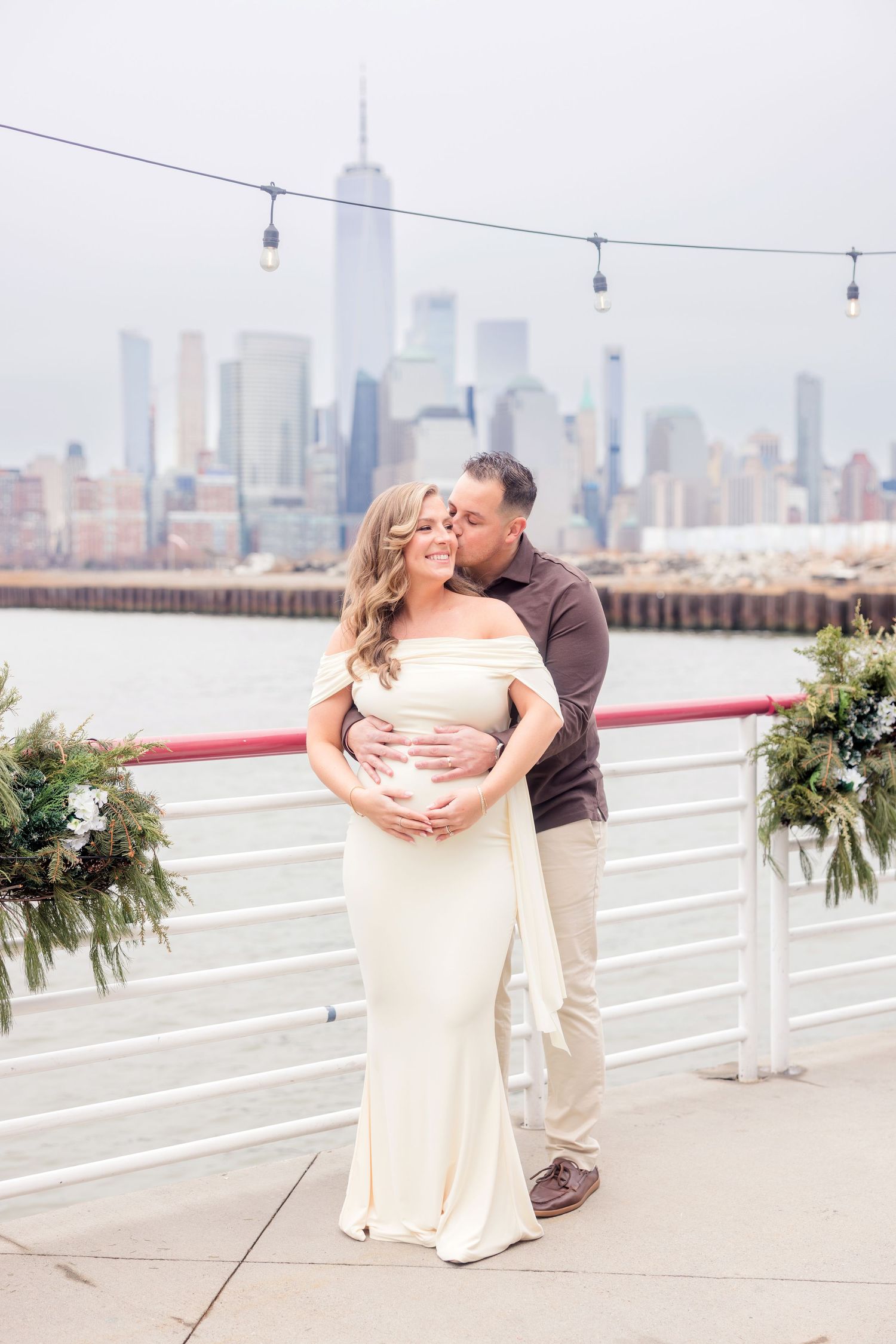 Couple embracing on decorated pier, man kisses pregnant woman in white gown with NYC skyline.