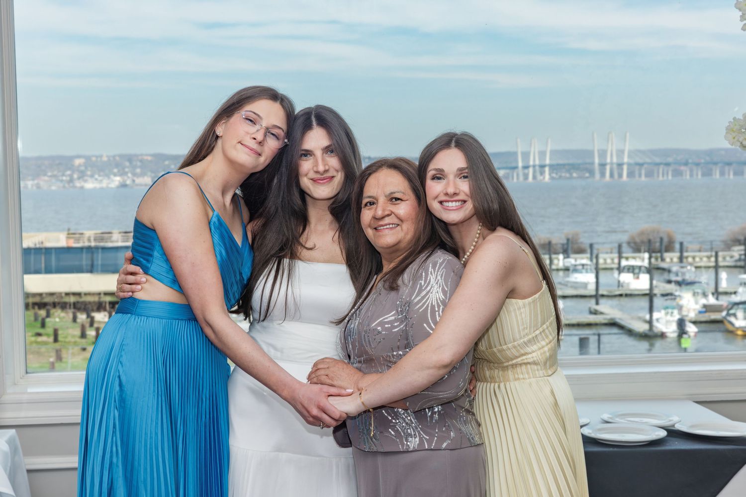 Four women in formal dresses posing with a scenic waterfront bridge view behind them.