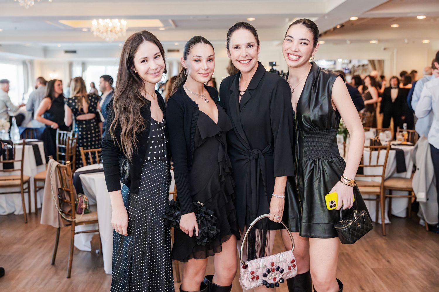 Four women in black dresses smiling together at an elegant social event with chandeliers.