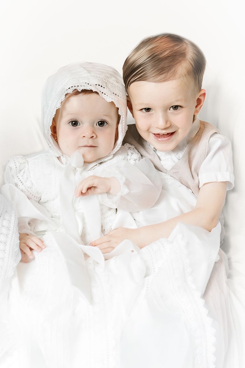 Two young children in white christening outfits, baby in bonnet held by smiling toddler, posed against white background.