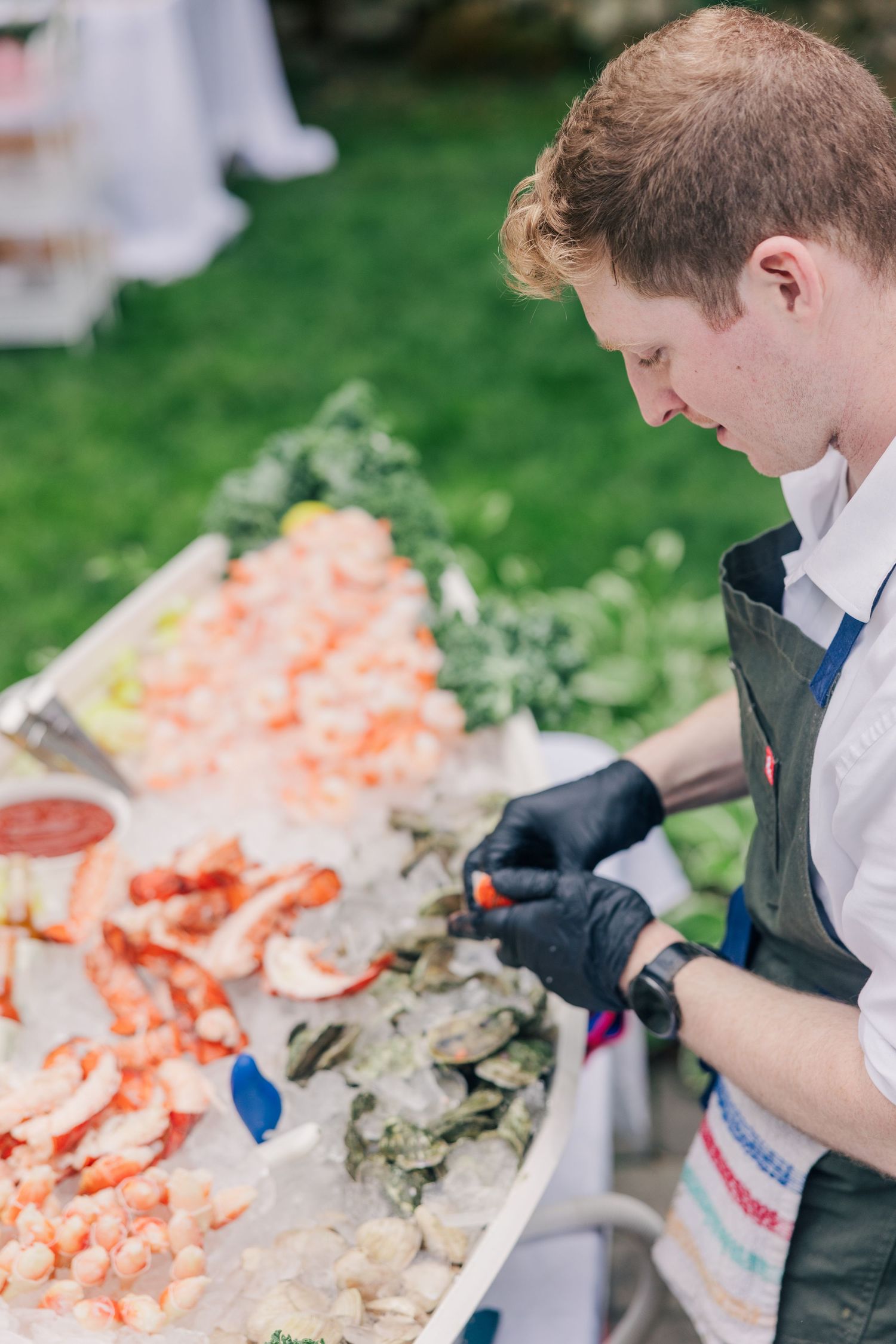 A catering staff member in black gloves arranges a seafood display with shrimp and oysters on ice at an outdoor event.
