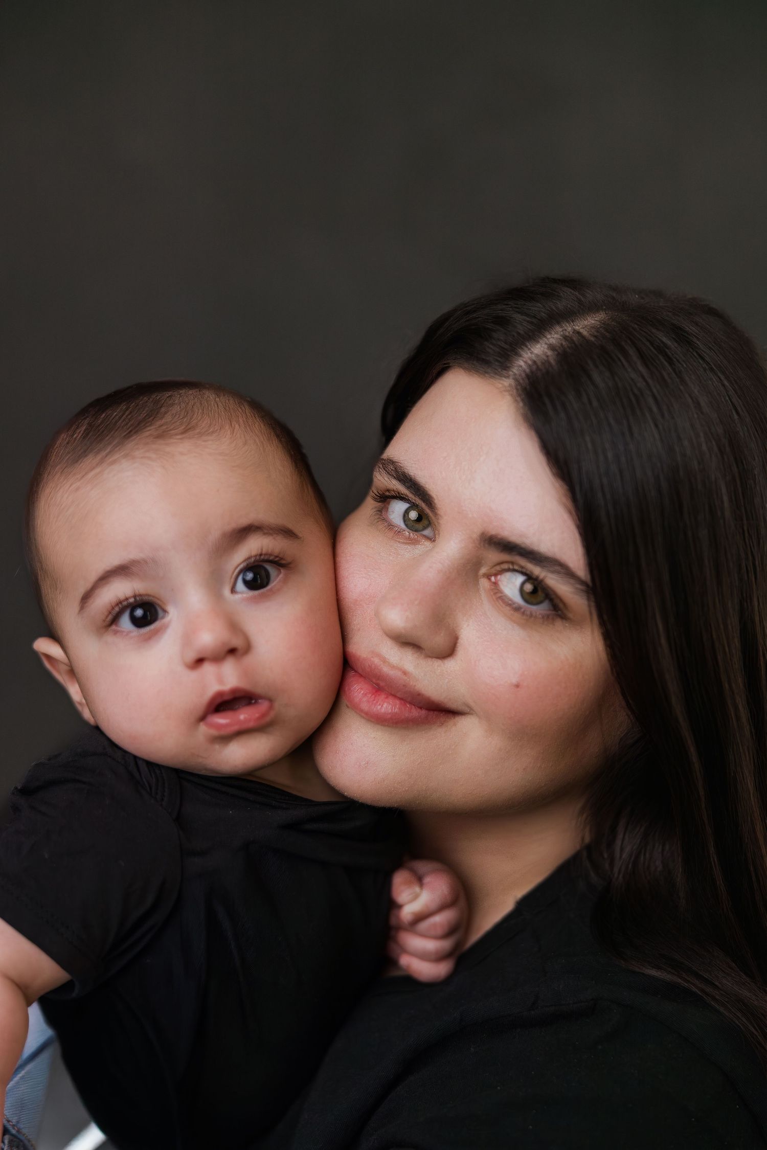 Young dark-haired mother in black shirt cheek-to-cheek with her wide-eyed baby against gray background.