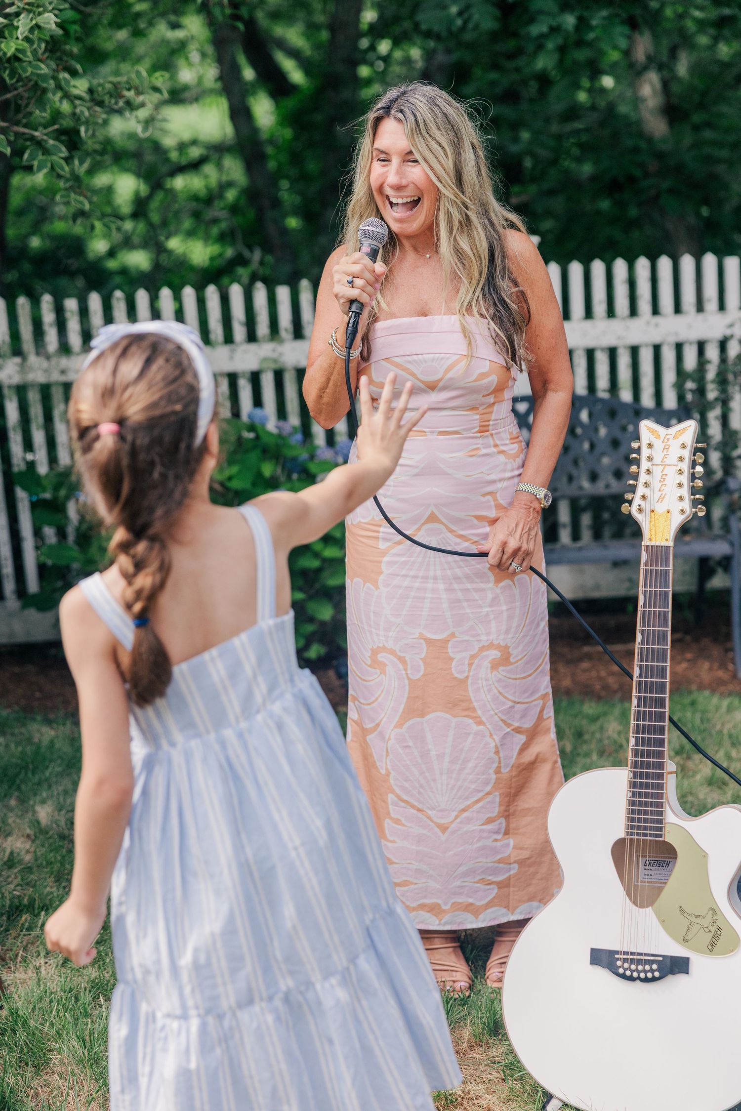 Woman in floral dress singing with microphone outdoors, young girl reaching toward her, white guitar nearby.