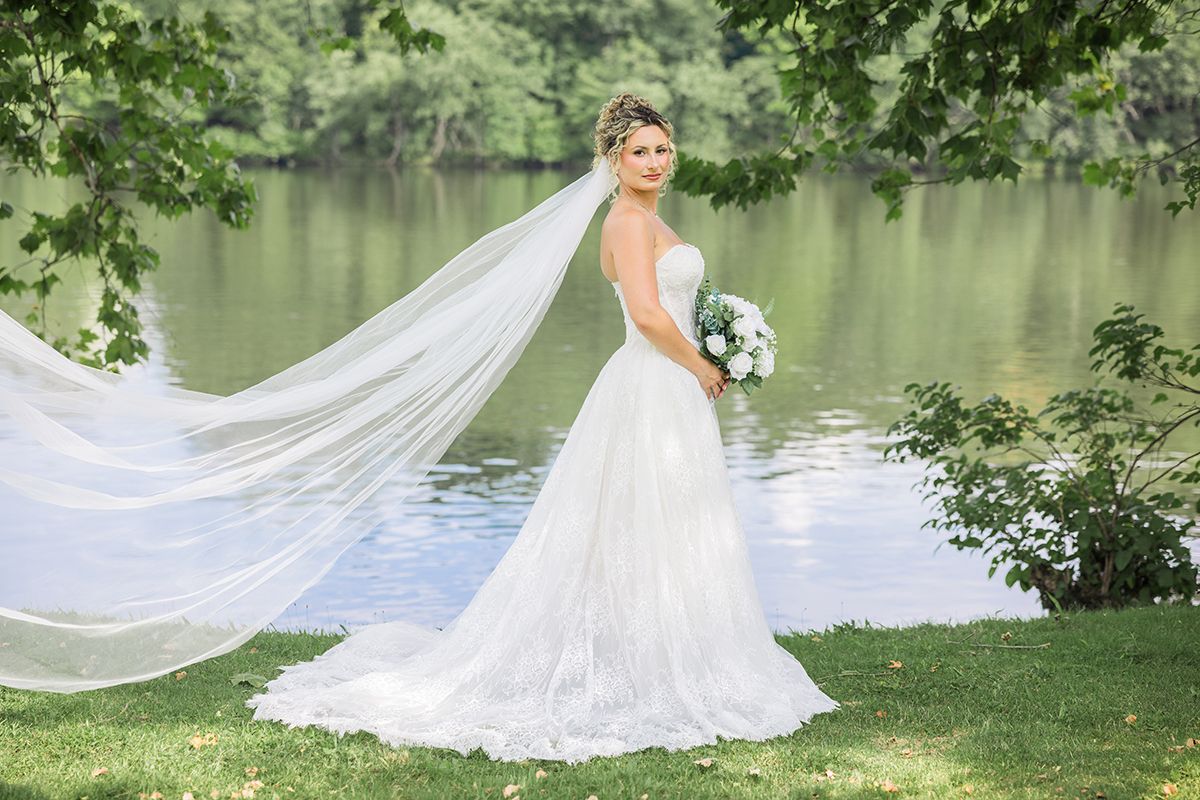 Bride in white gown with flowing veil stands by a serene lake surrounded by lush green trees holding a bouquet.