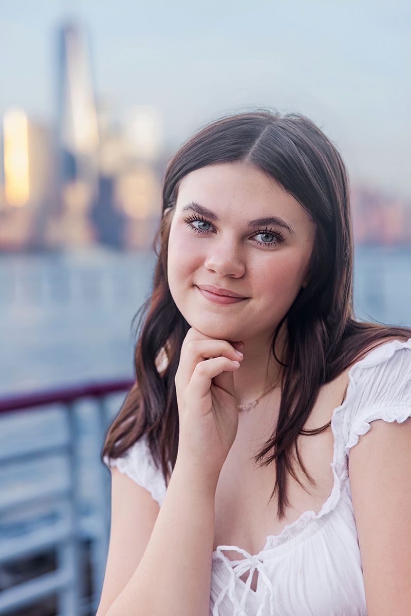 Young woman with blue eyes in white dress poses thoughtfully by waterfront with city skyline at dusk.