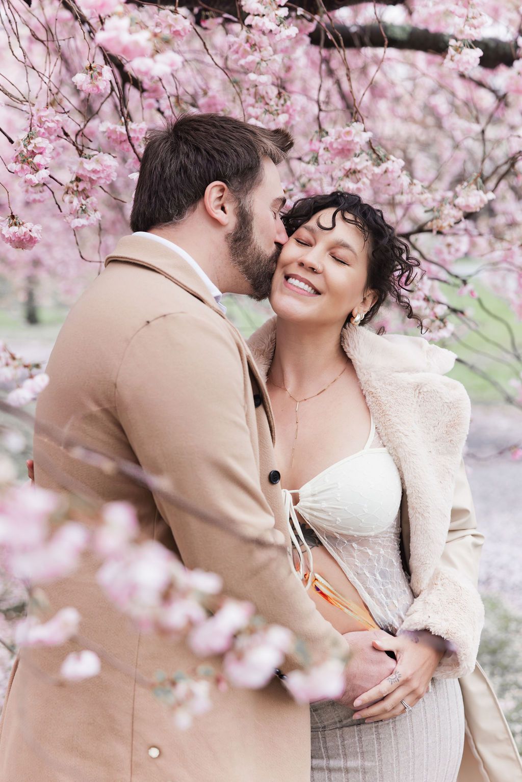 Man kisses smiling pregnant woman's cheek under blooming pink cherry blossom trees in spring.