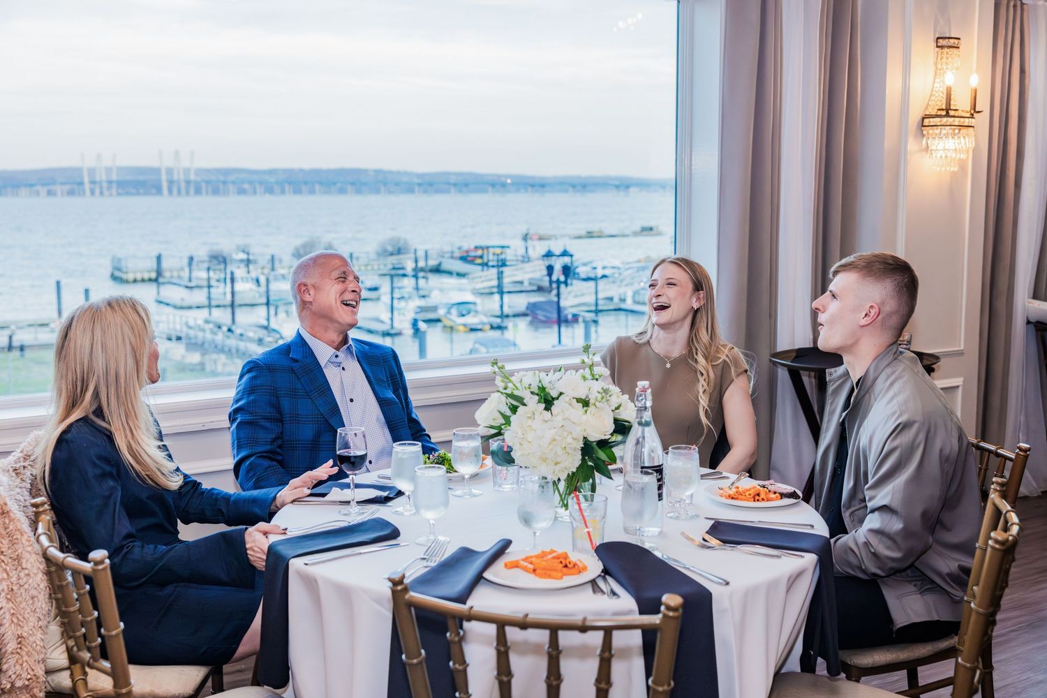 Four guests laugh and enjoy conversation at a waterfront restaurant with scenic harbor views.