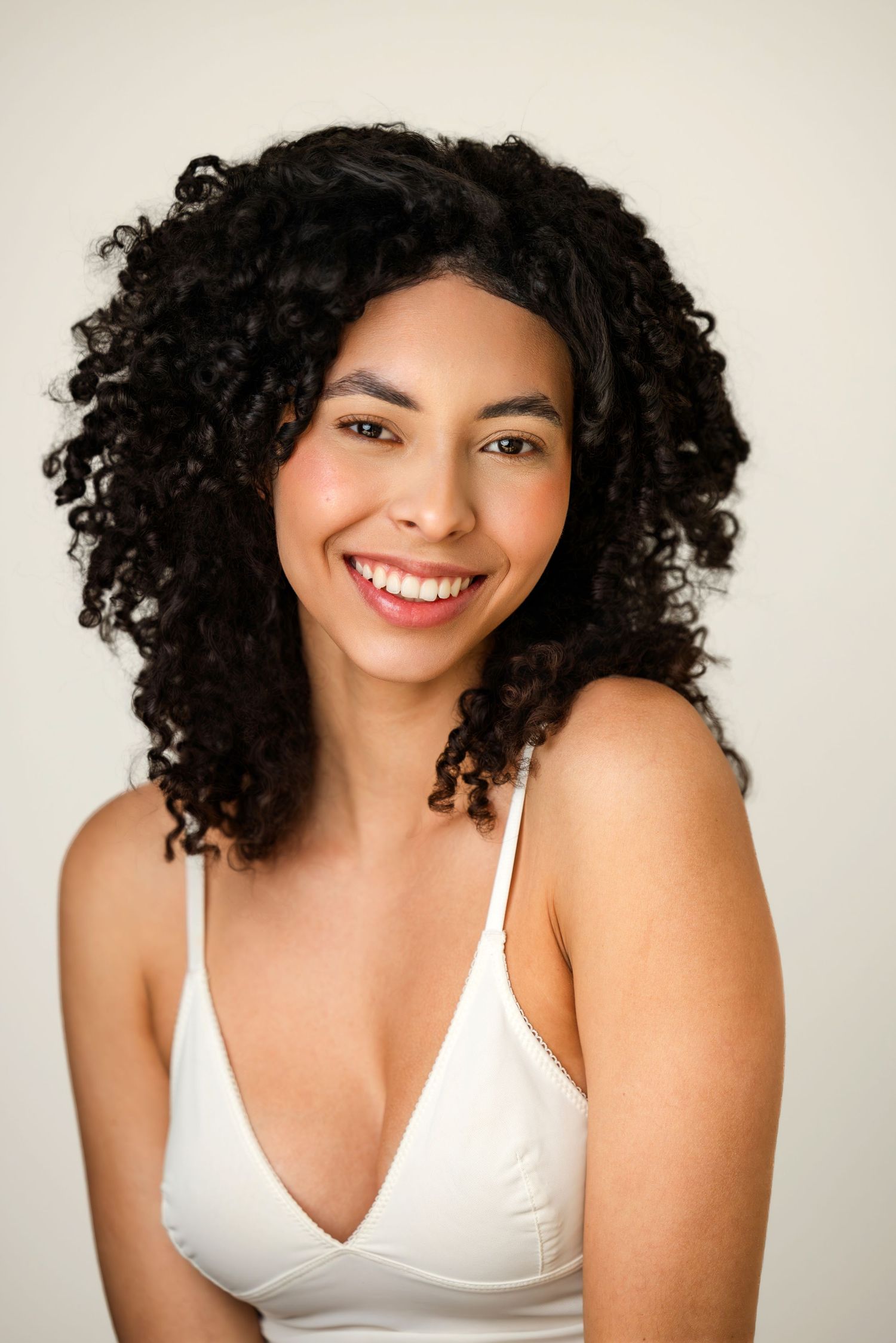 Smiling woman with curly black hair wearing white bralette against neutral background.