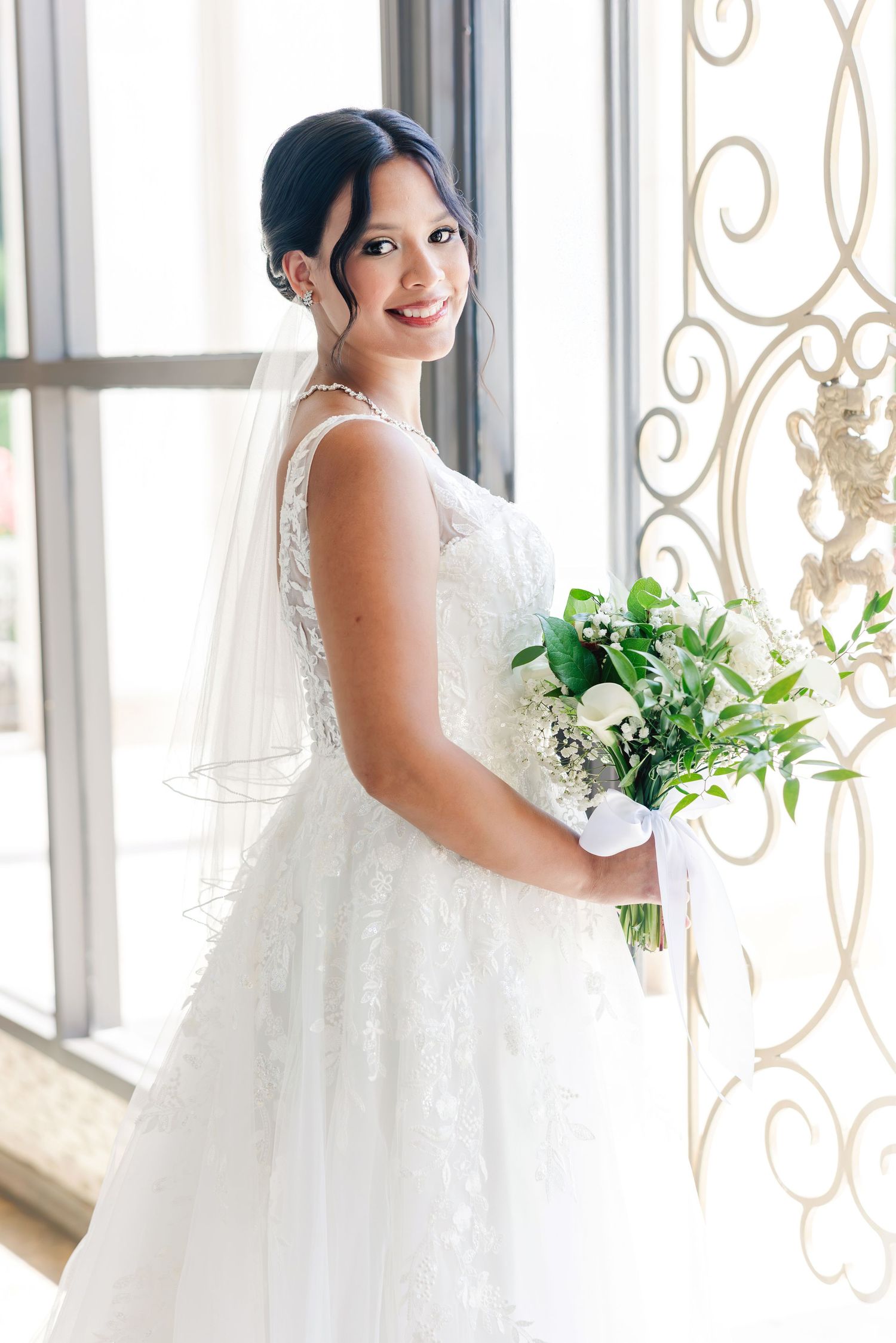 Smiling bride in lace wedding dress holding green and white bouquet standing by decorative window.