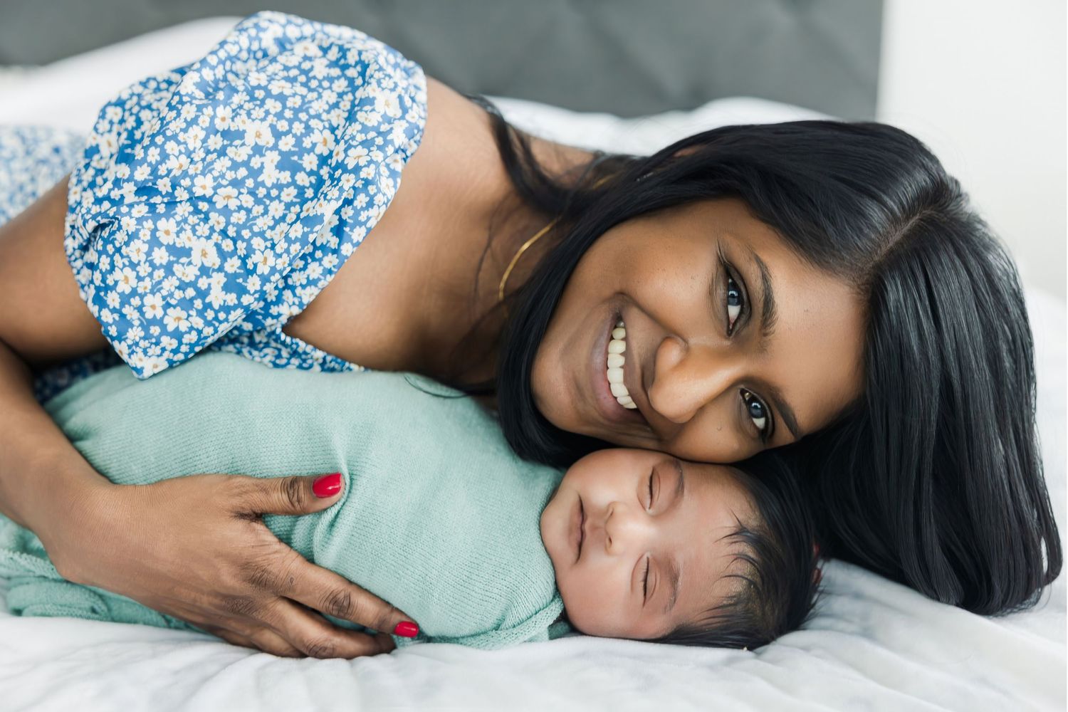 Smiling mother lying on white bed cheek-to-cheek with her newborn baby wrapped in green swaddle.