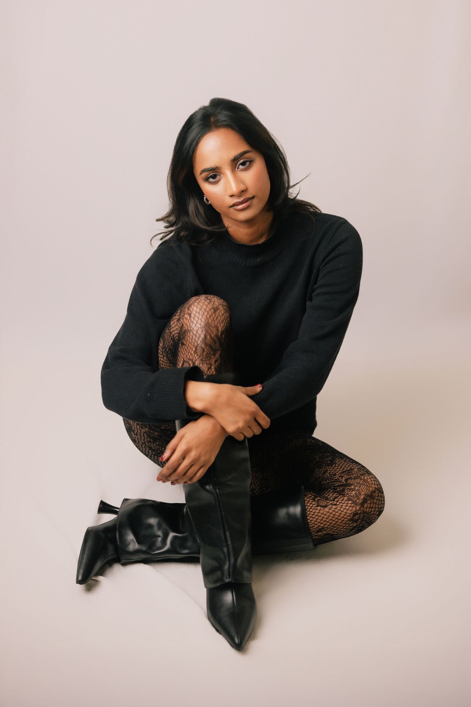 Woman in black sweater, lace tights, and boots sitting on floor against light background.