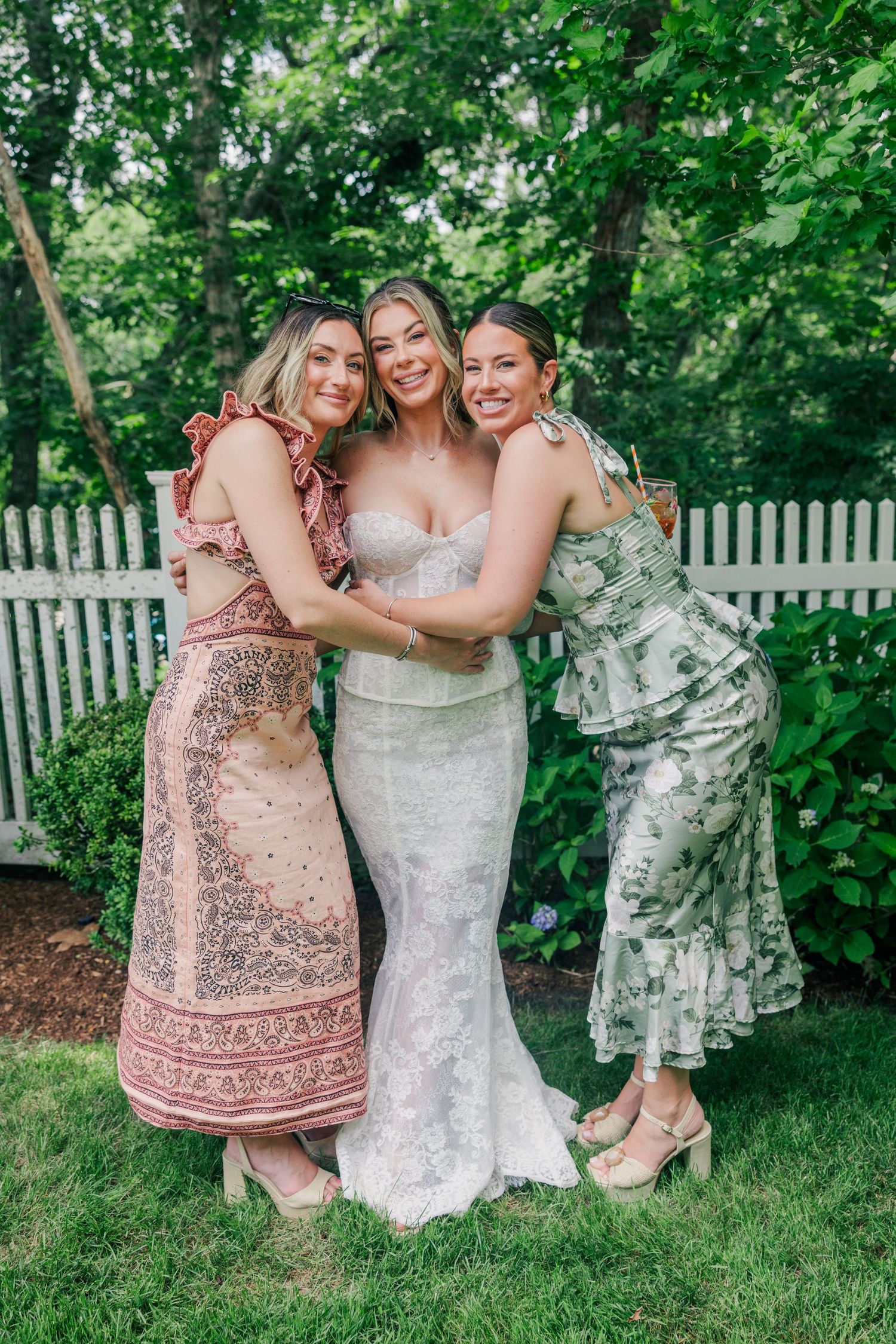 A bride in a white lace gown hugging two friends in floral dresses outdoors near a white picket fence.