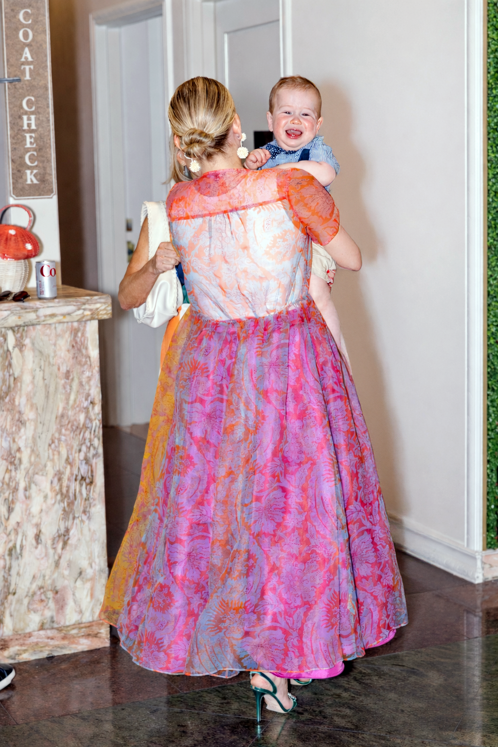 Woman in colorful floral gown holds laughing baby at coat check area of formal event.