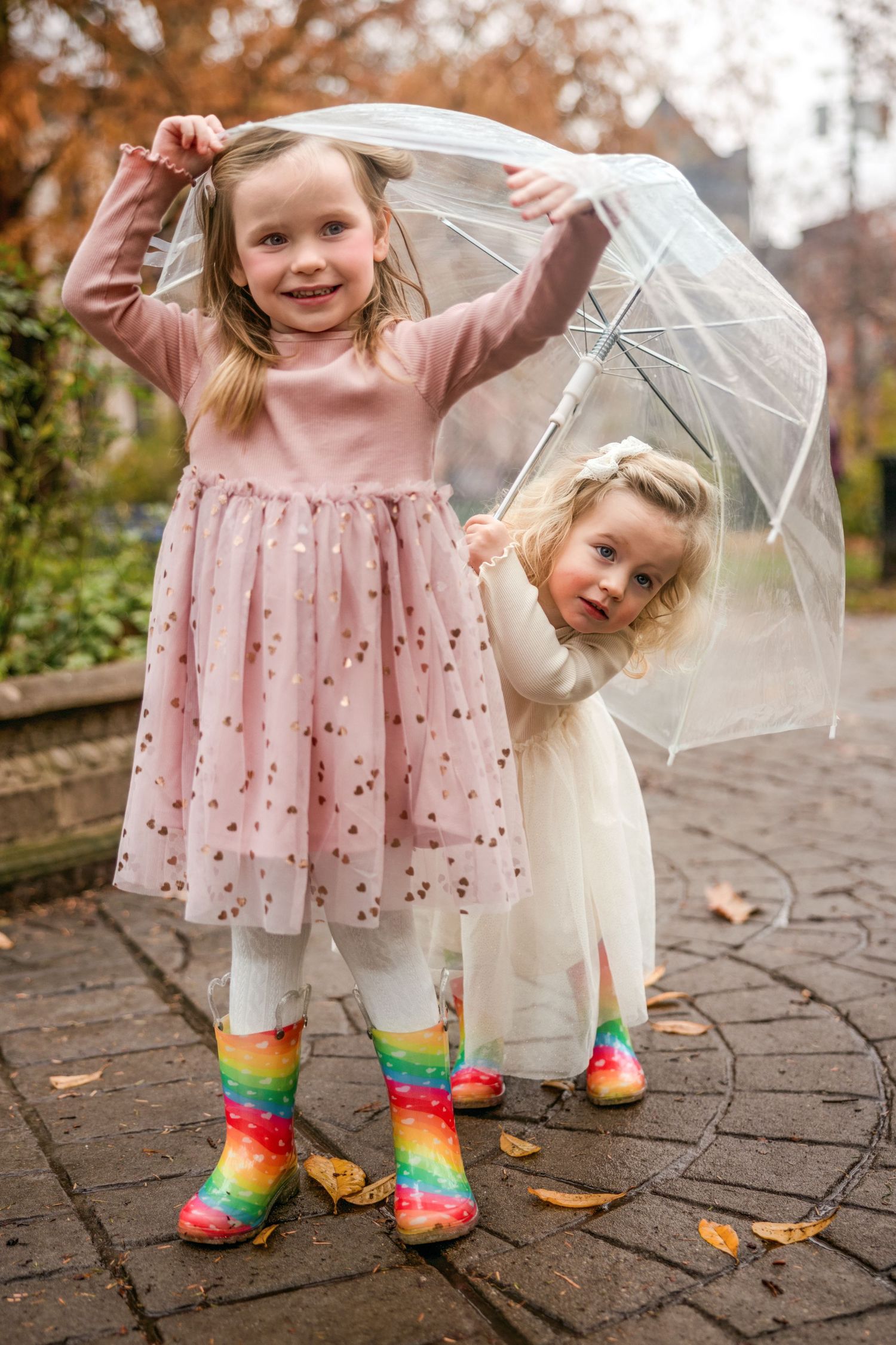 Two little girls sharing a clear umbrella outdoors, one in pink tutu dress with rainbow rain boots.