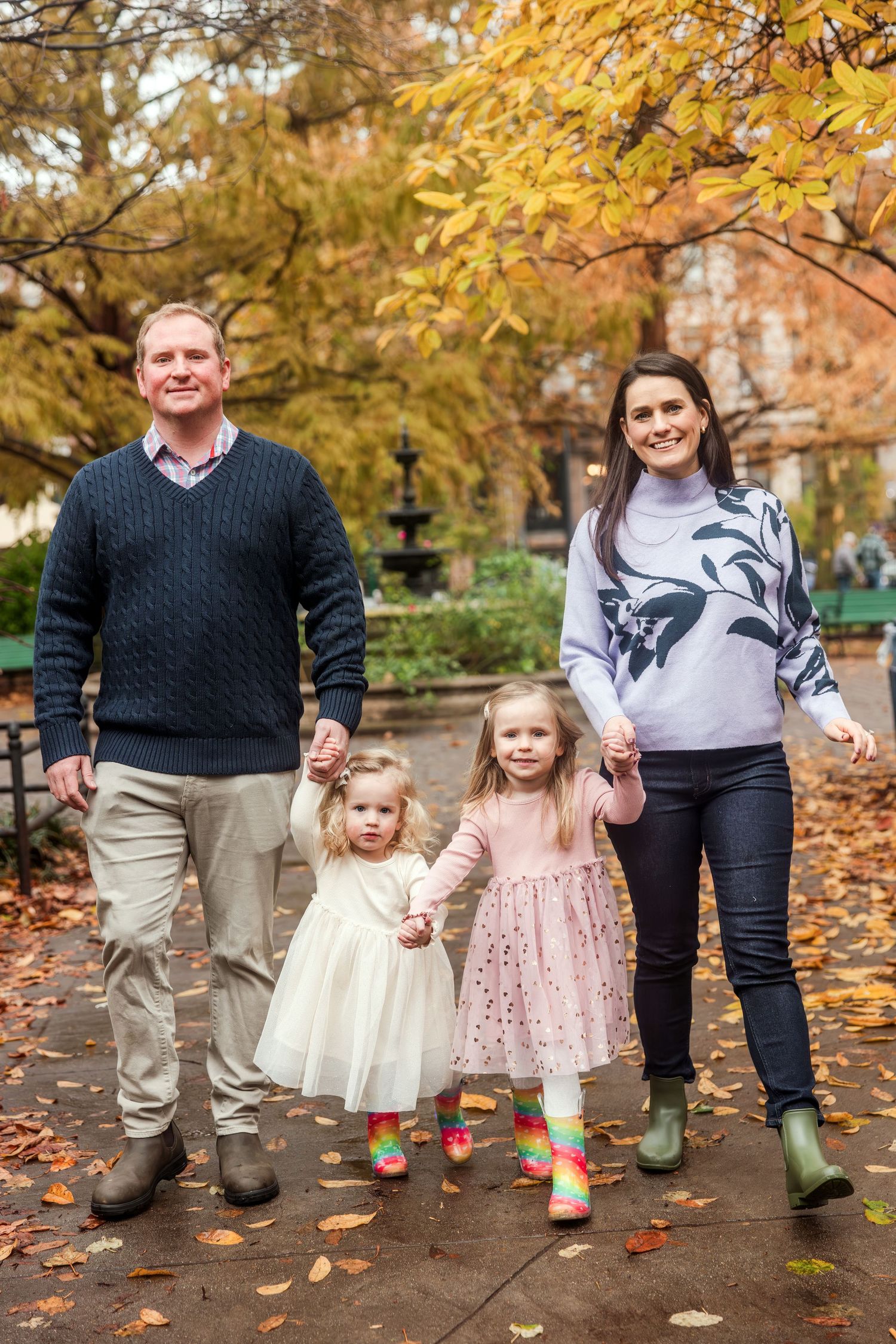 Family of four holding hands walking through colorful autumn park, two young daughters in festive dresses.