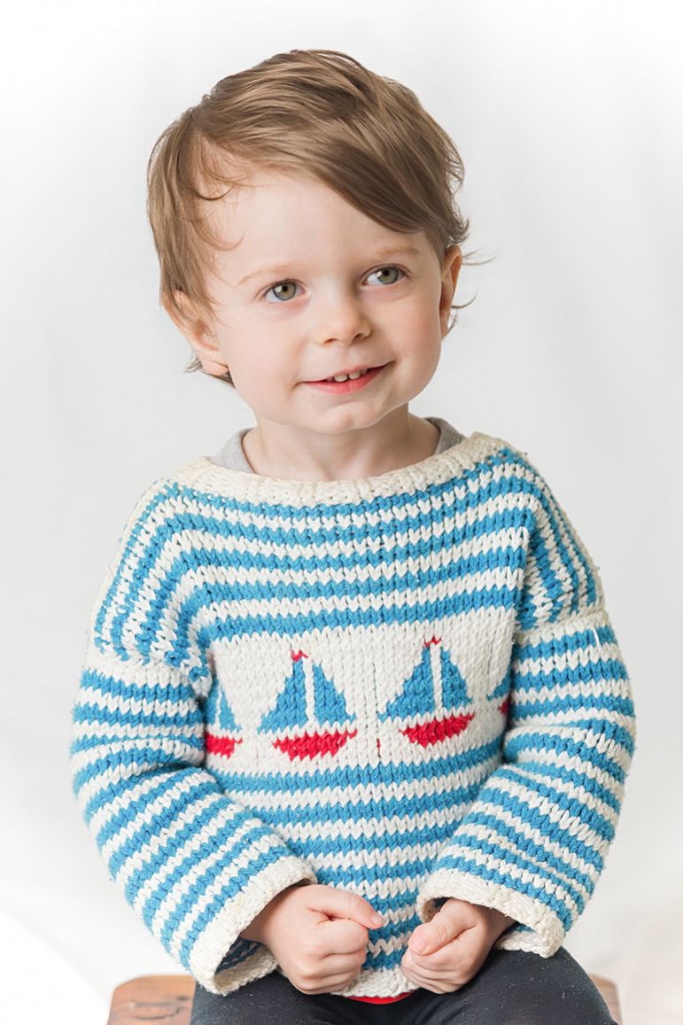 Smiling toddler boy wearing a blue and white striped knit sweater with sailboat designs, sitting against a white background.