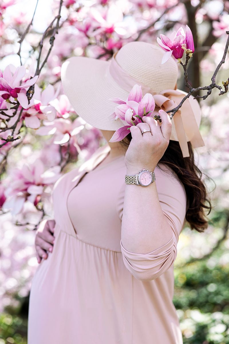 Pregnant woman in pink dress smells magnolia blossoms wearing wide-brim hat among pink flowers.