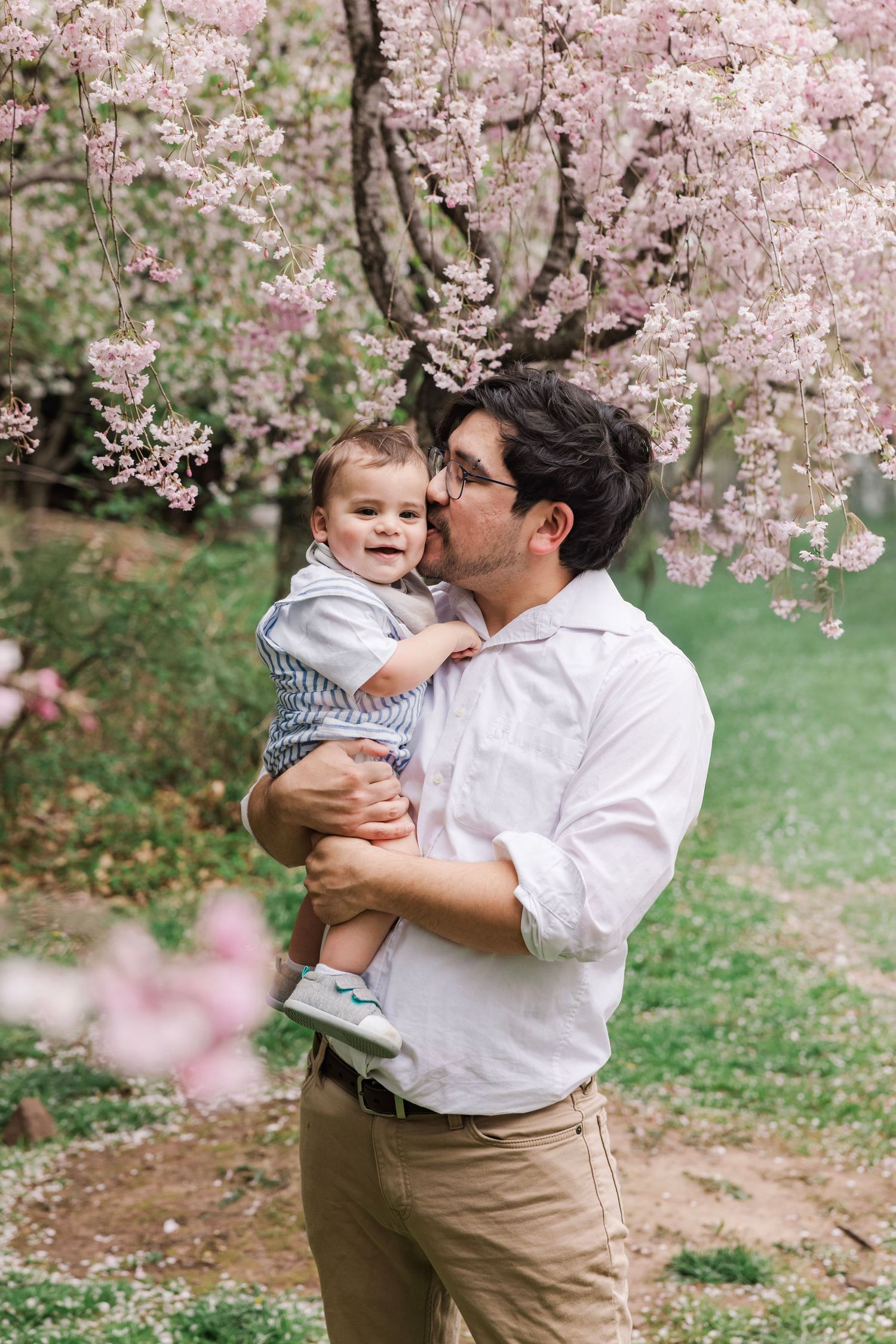 A loving father kisses his smiling baby boy while standing under pink cherry blossom trees in spring.