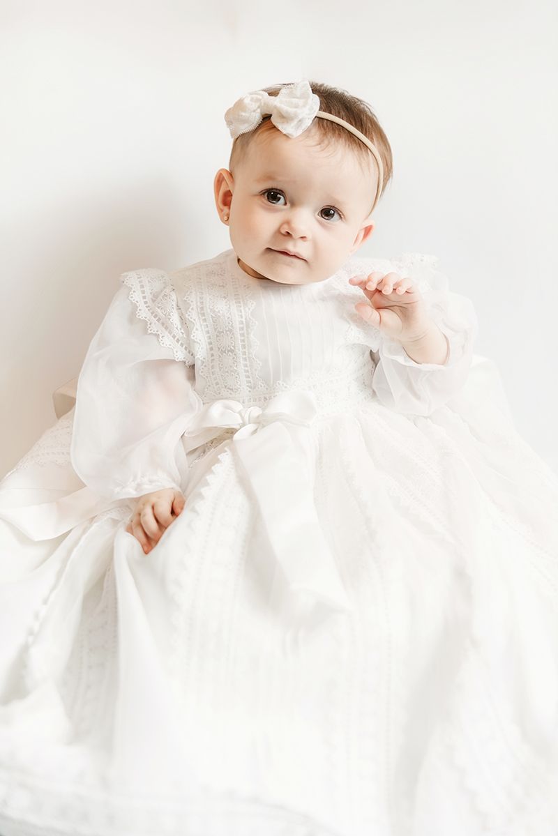 Adorable baby girl in white christening gown with bow headband, sitting against white background.