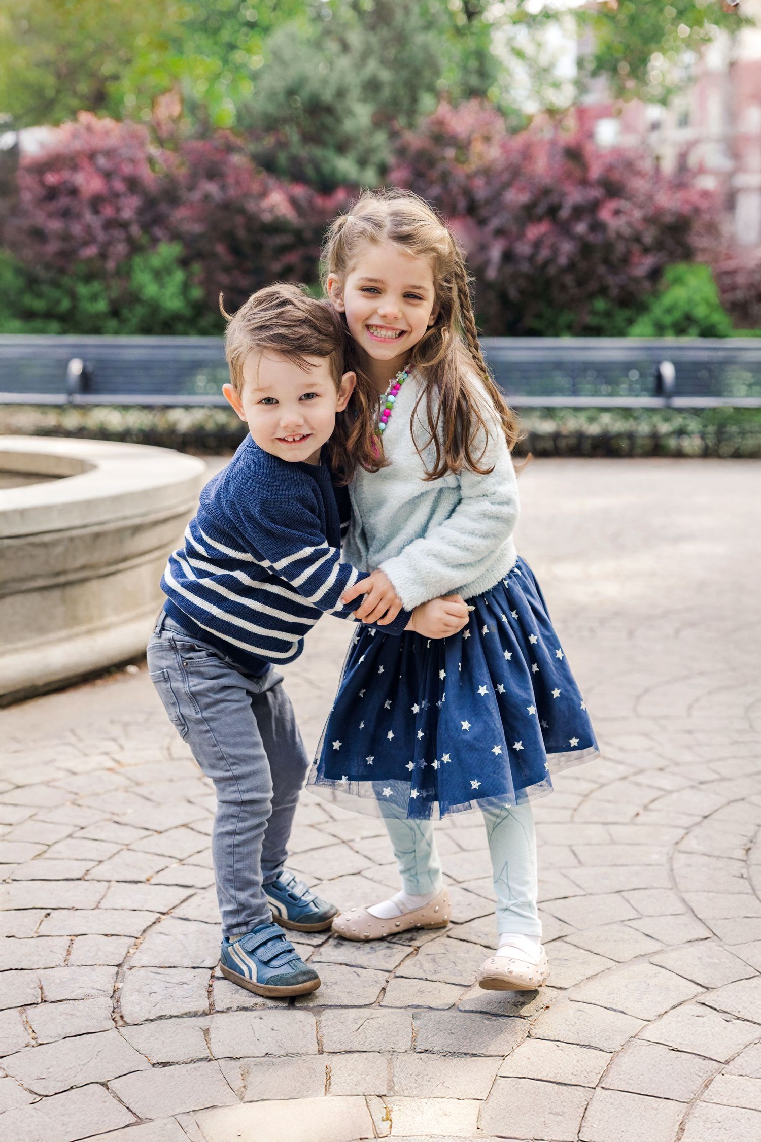 Two young children hugging and smiling in a park with colorful foliage in the background.