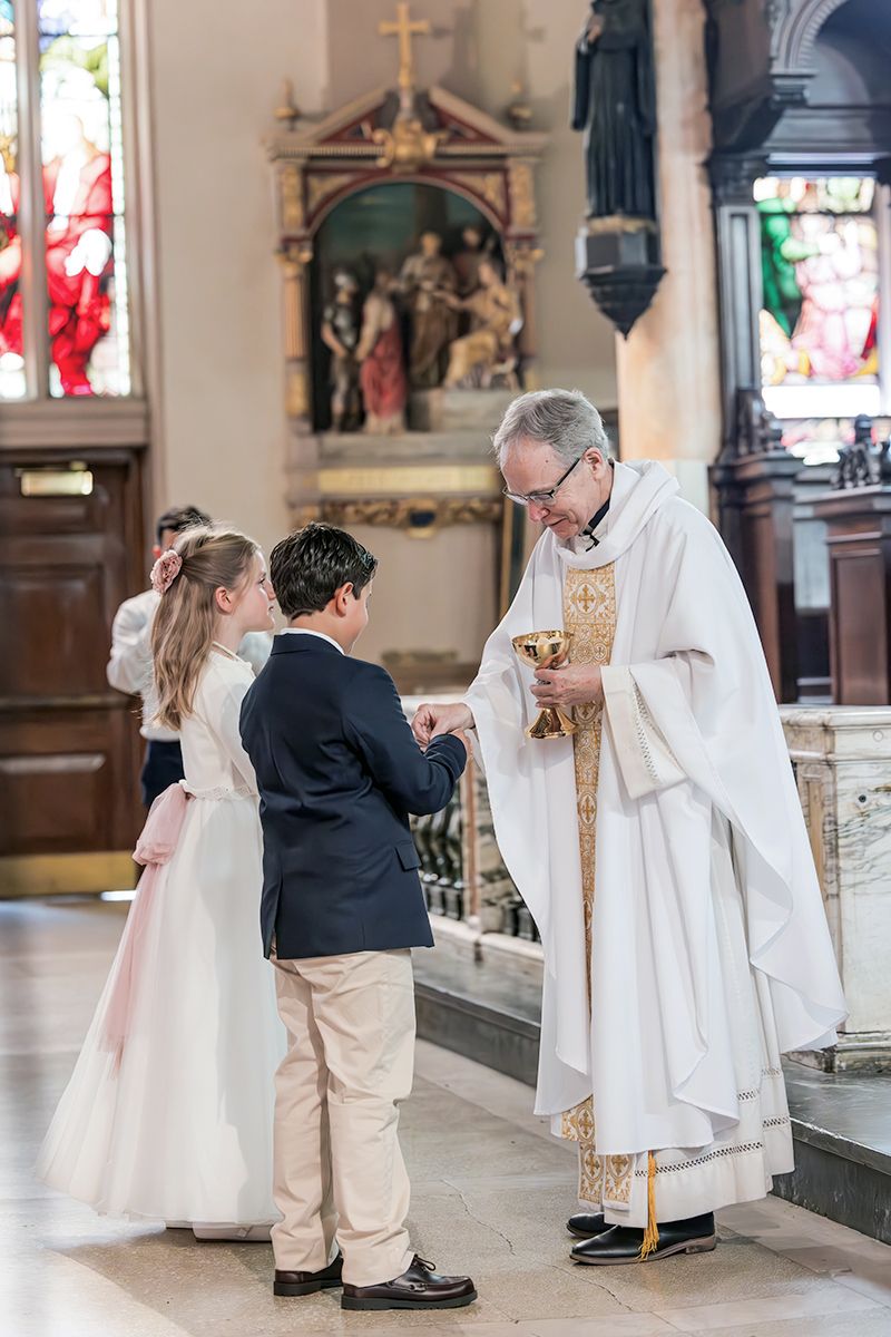 Priest giving First Communion to children in a decorated Catholic church with stained glass windows.