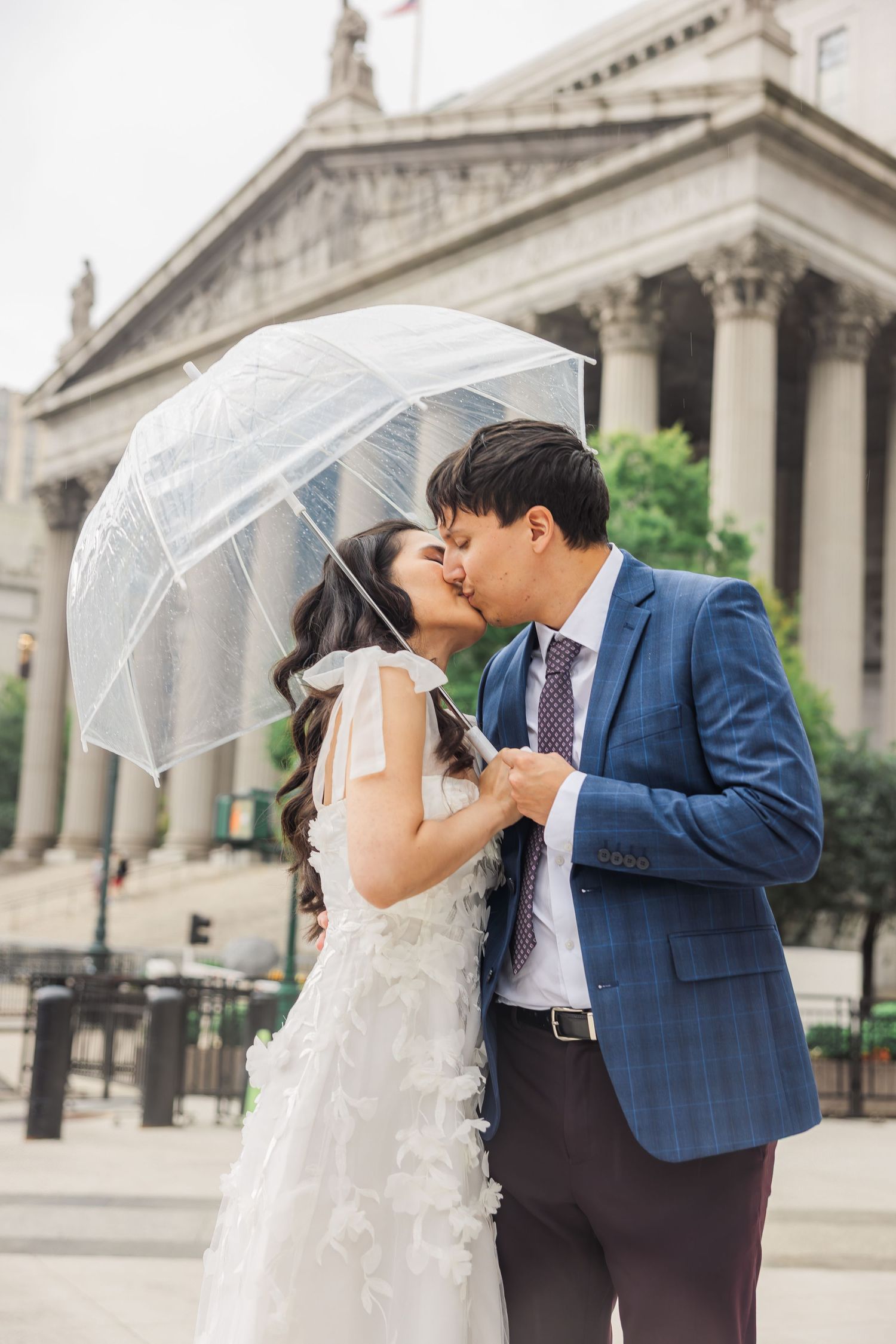 A bride and groom share a kiss under a clear umbrella outside a grand neoclassical building.