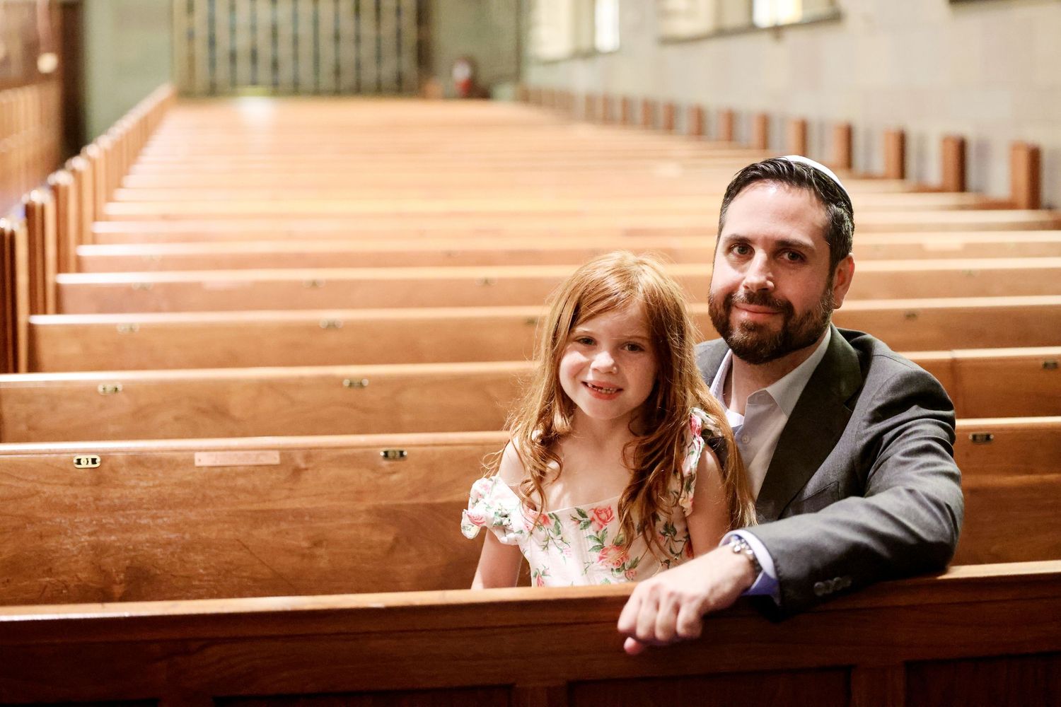 Bearded father in suit sits with young red-haired daughter in church pew smiling.