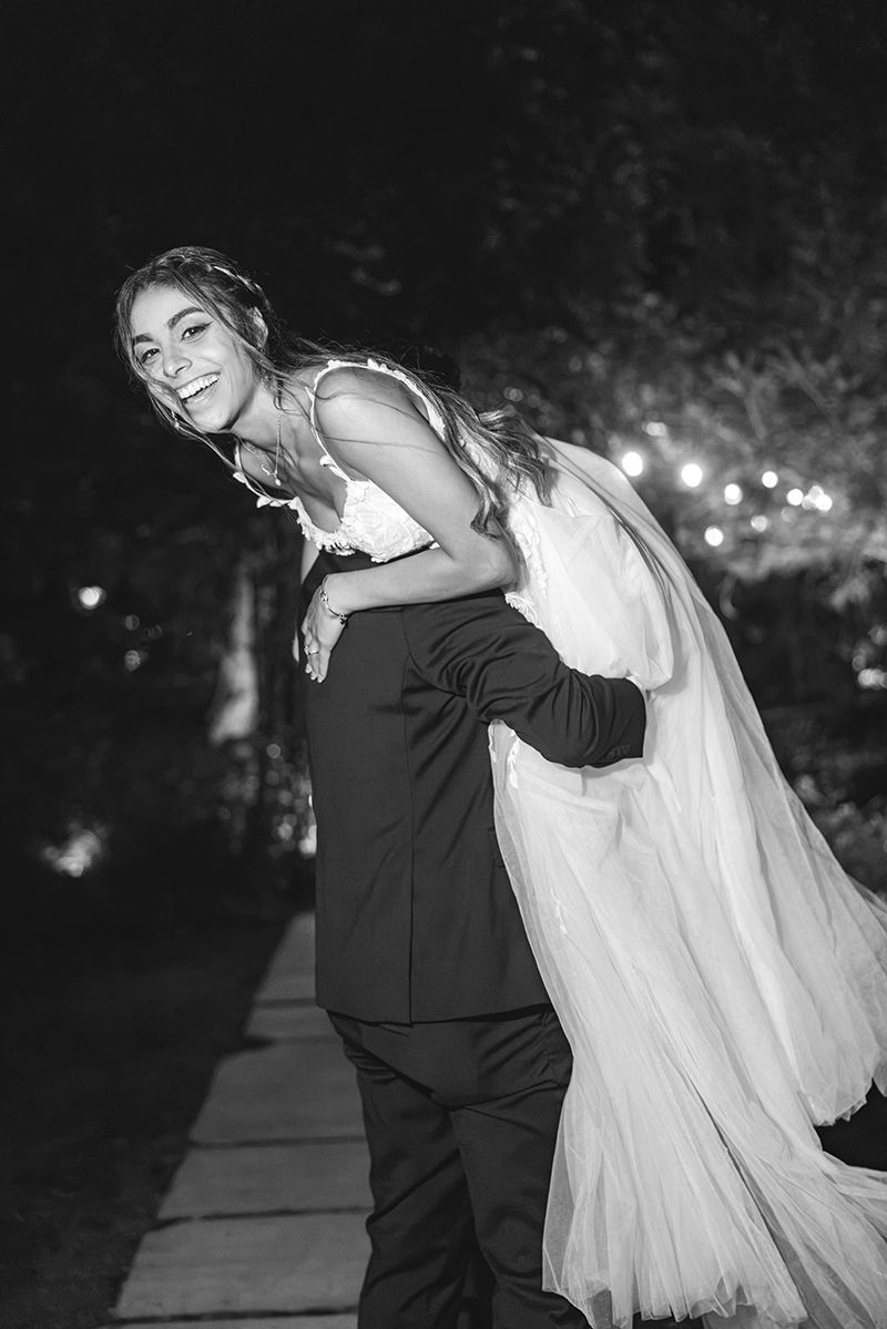 Black and white photo of a groom carrying his bride in a wedding gown outdoors at night, both smiling joyfully.