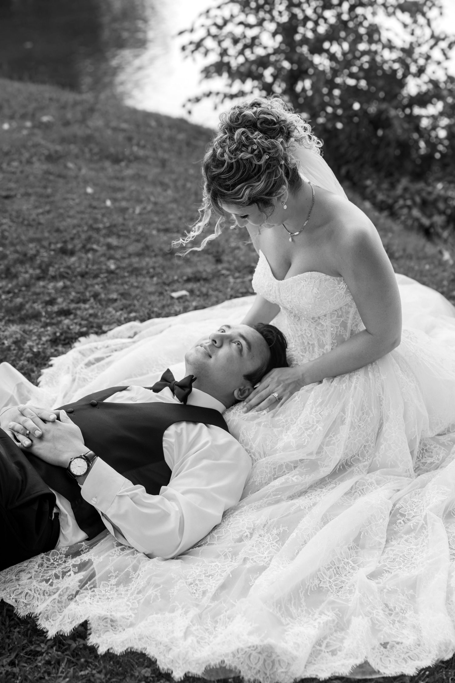 Black and white wedding photo of bride and groom relaxing on grass, groom lying down, bride in lace gown smiling at him.