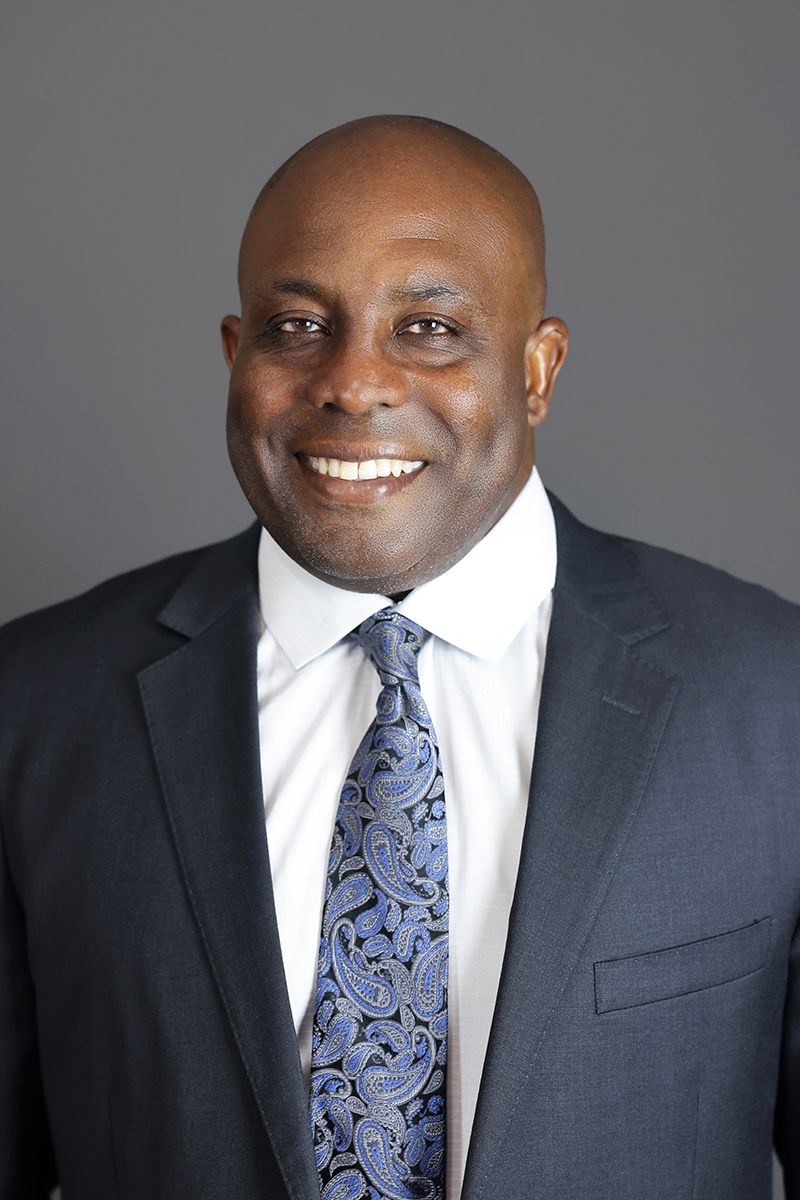 Professional headshot of a smiling bald man in a dark suit and blue paisley tie against a gray background.