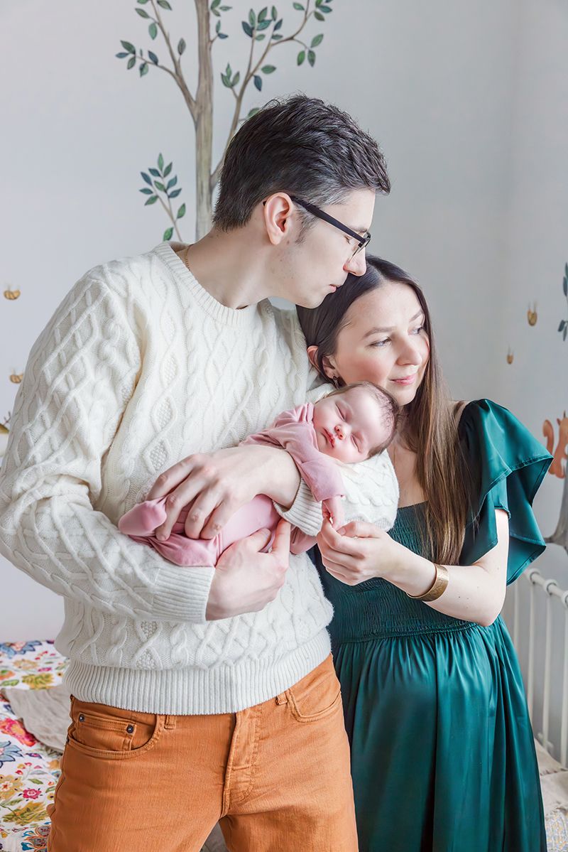 Young parents in nursery tenderly holding sleeping newborn baby in woodland-themed room.