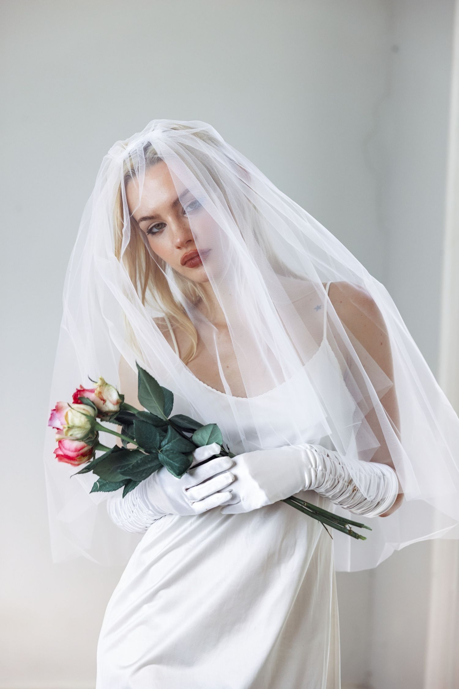 Bride in white gown, veil, and gloves holding a bouquet of roses in a soft, ethereal bridal portrait.