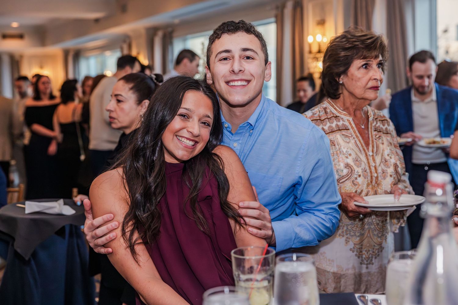 A young man and woman smile and embrace while seated at a table during a formal event.