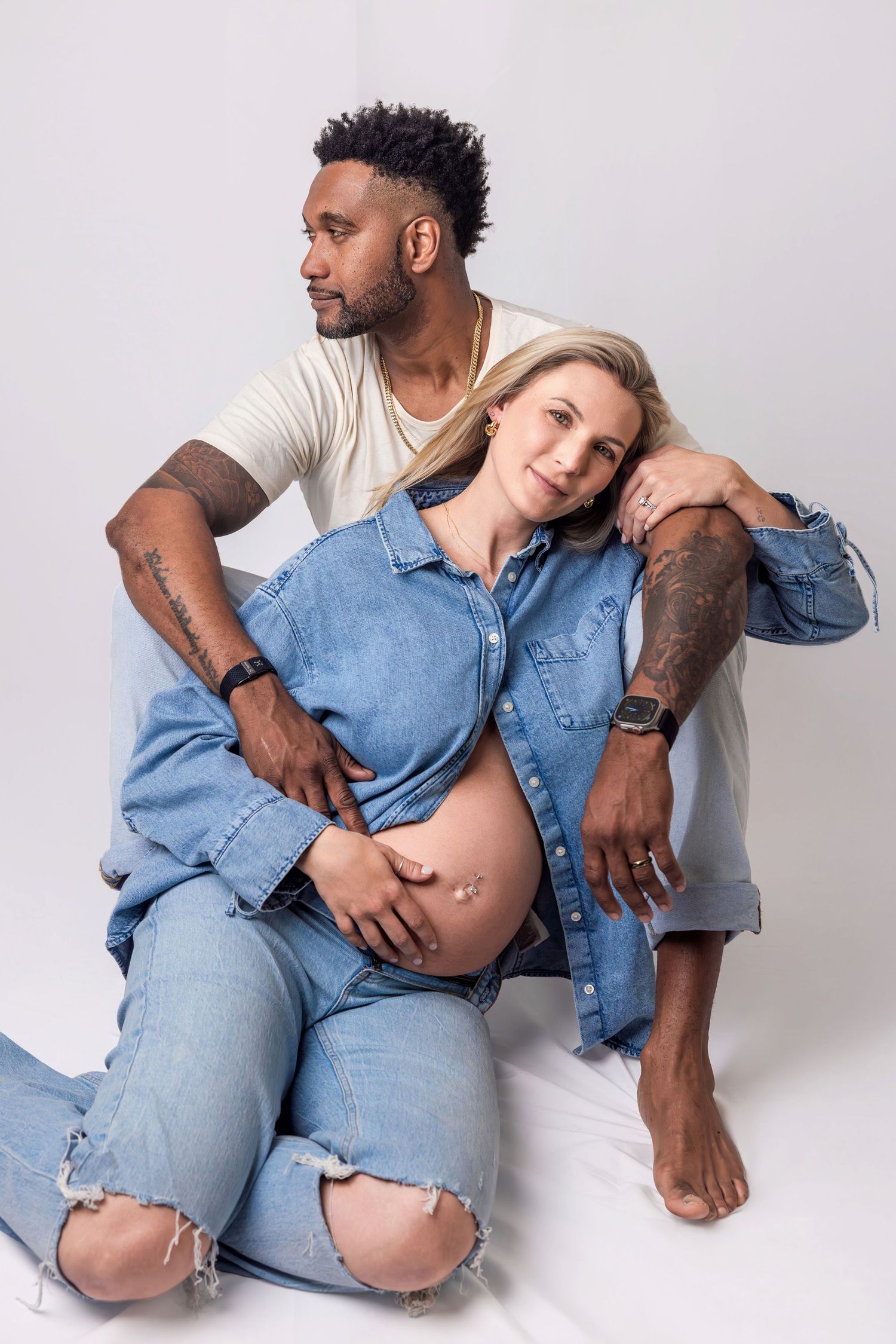 Interracial couple sits on floor together cradling pregnant womans bare belly.