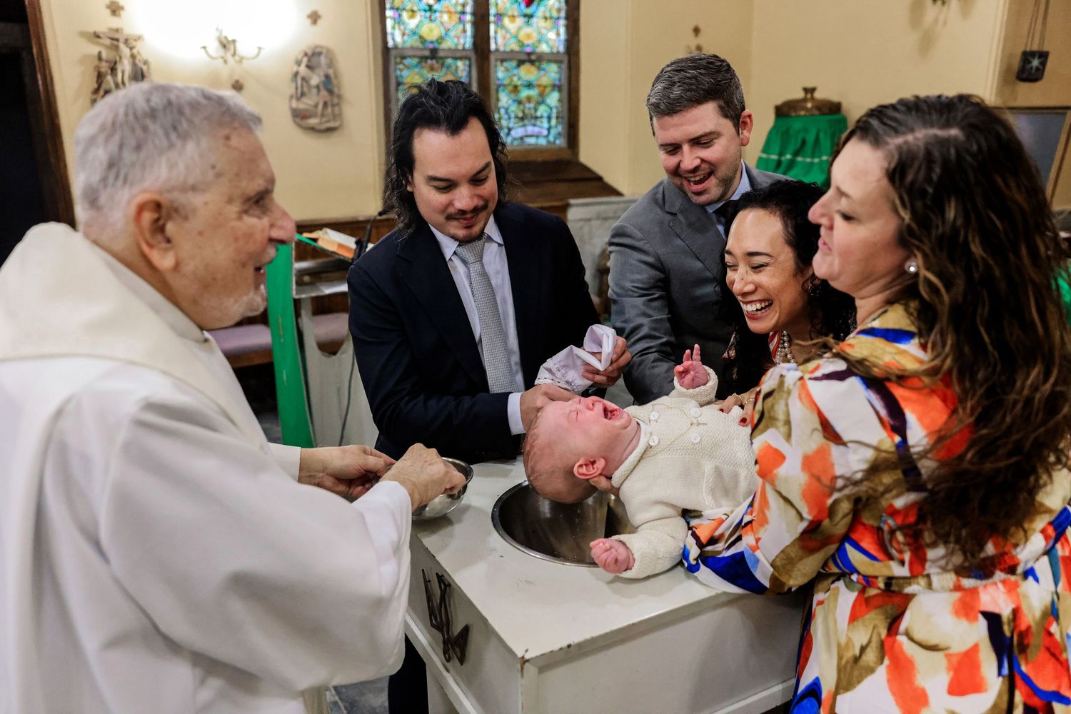 Priest performs infant baptism with family gathered around the baptismal font.