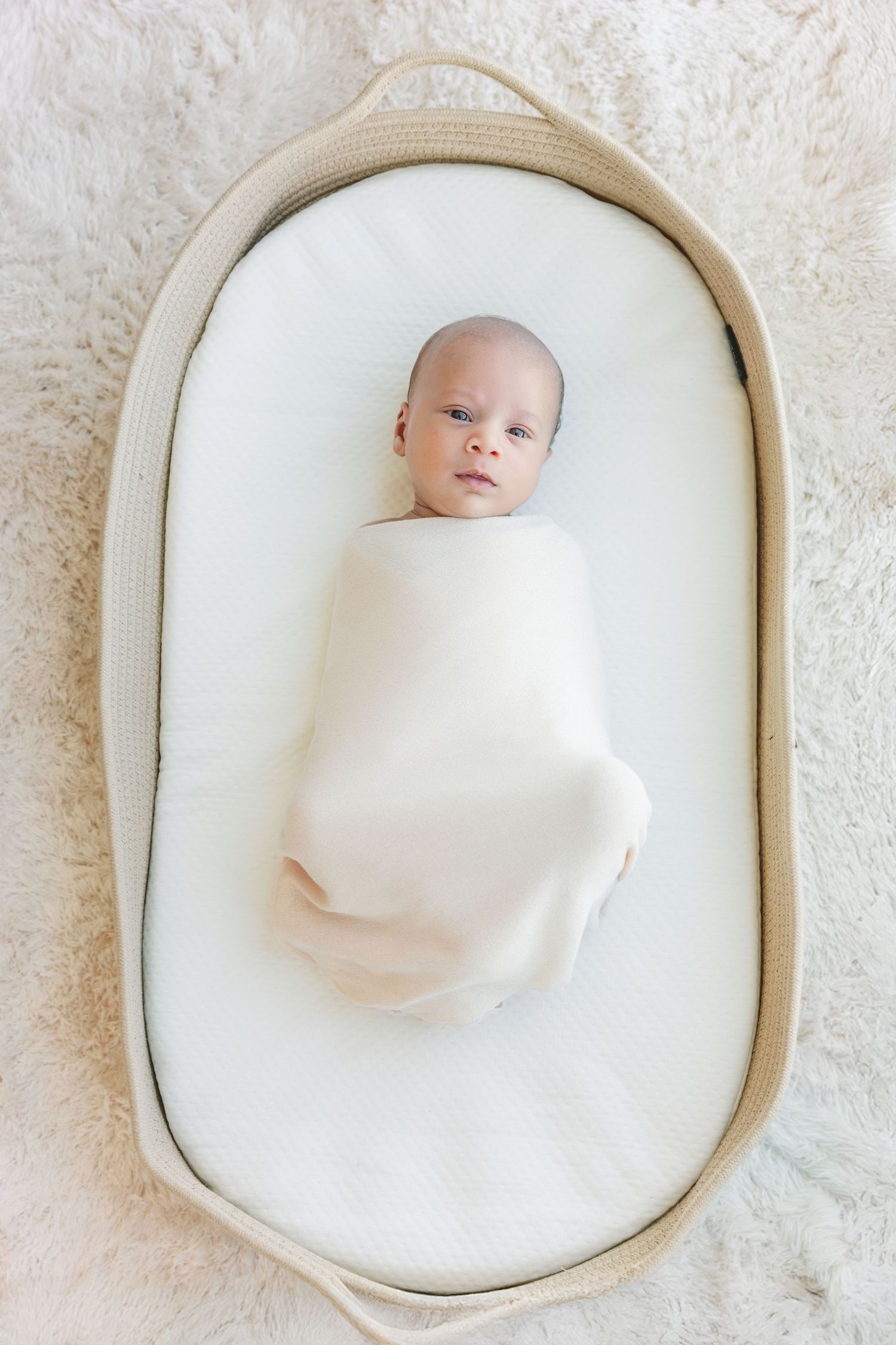 Newborn baby swaddled in white wrap lying in an oval wicker bassinet on a soft cream fur surface.