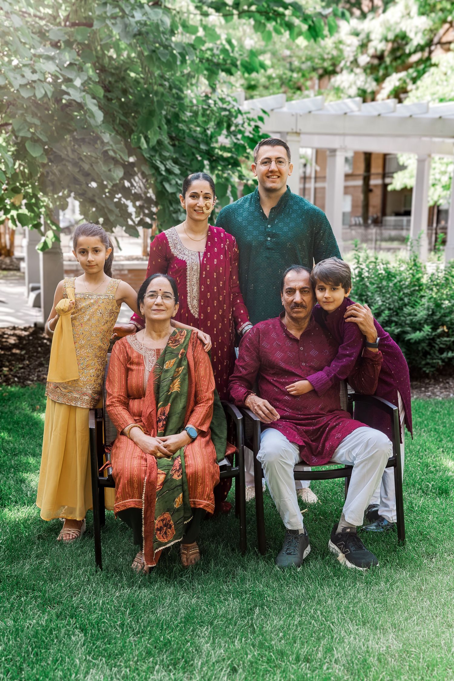 Multi-generational South Asian family posing together outdoors in traditional colorful attire on a lush green lawn.