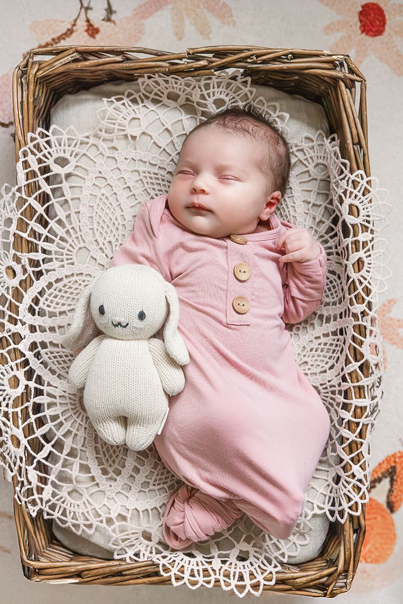 Sleeping newborn baby in pink outfit with stuffed bunny toy in wicker basket with white lace doily.