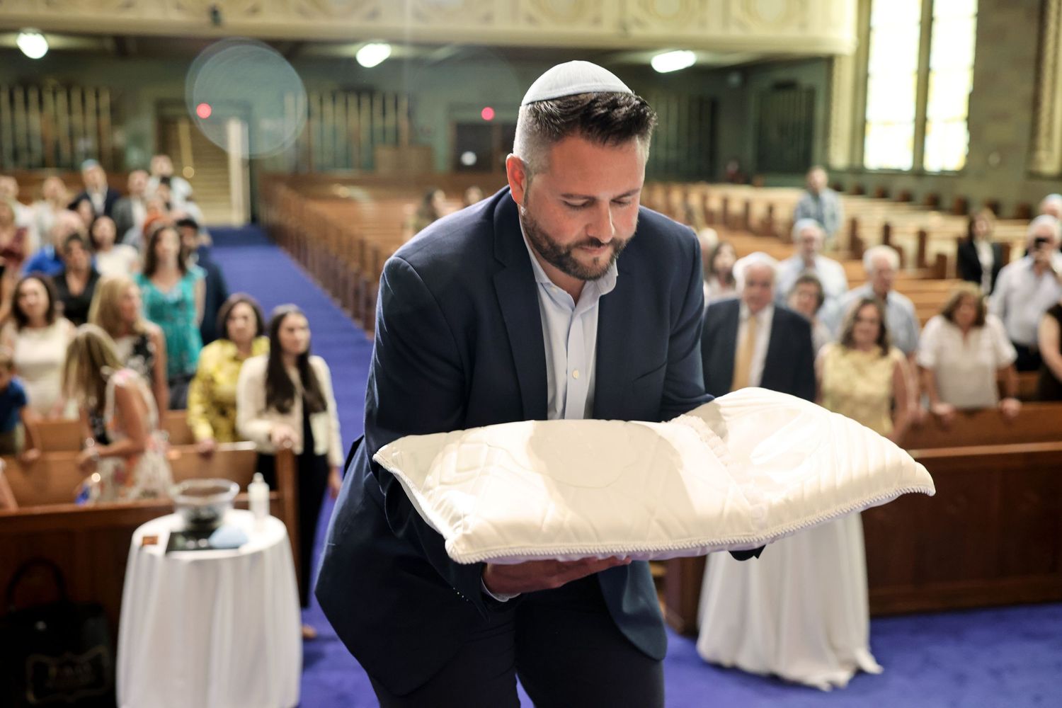 Man wearing kippah carefully carries baby on ceremonial pillow down synagogue aisle.
