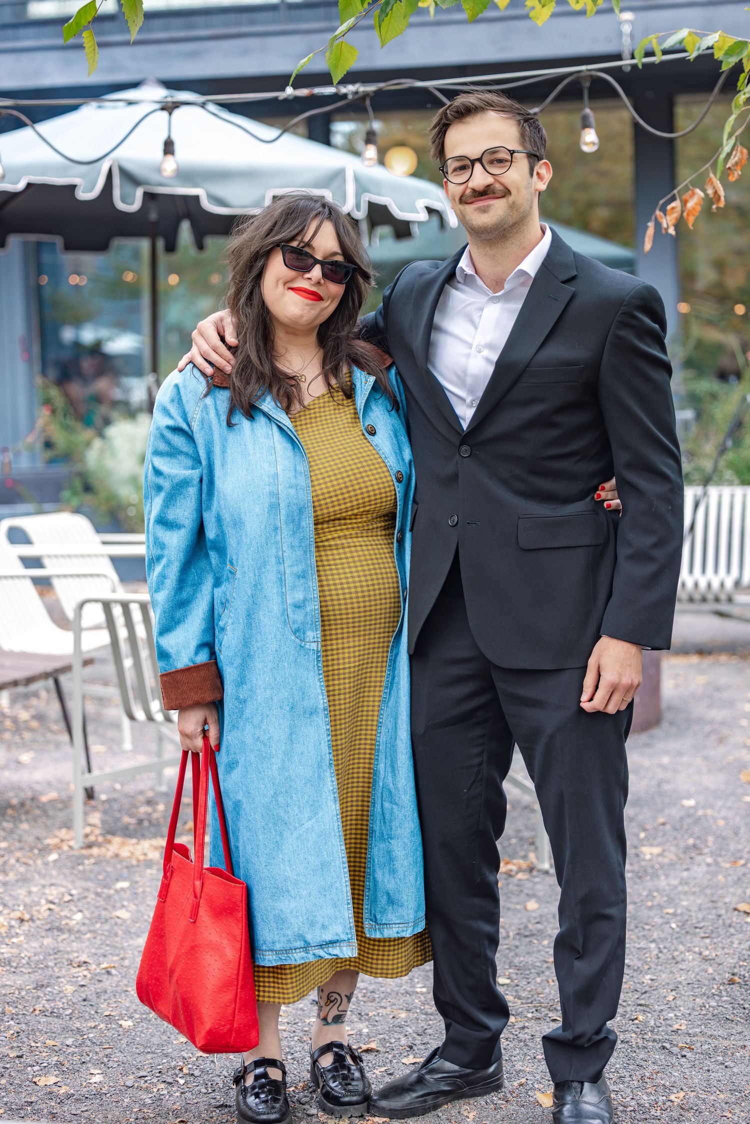 Pregnant woman in olive dress and blue coat with man in black suit posing outdoors.
