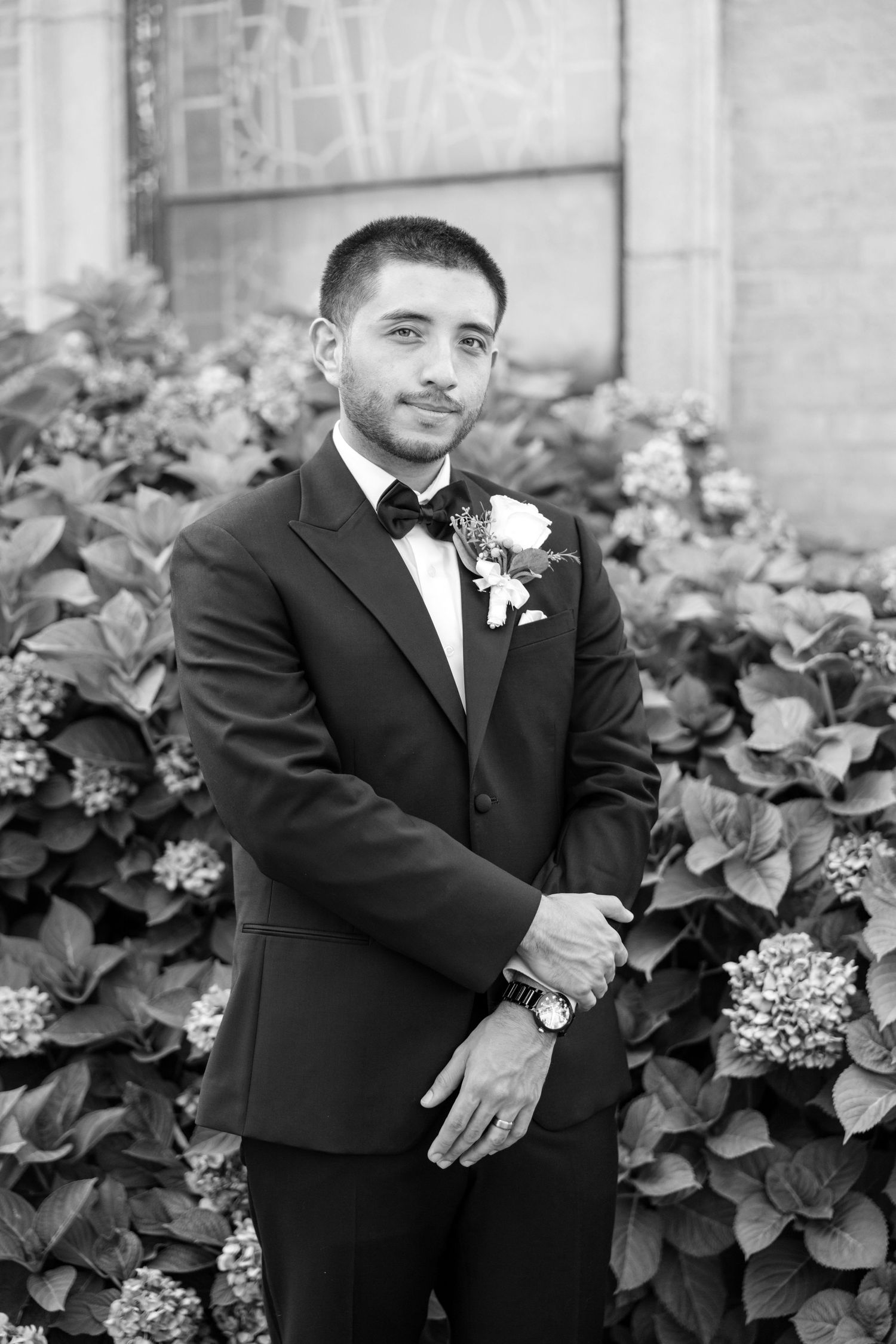 Groom in black tuxedo with bow tie and boutonniere poses among lush hydrangeas in elegant black and white wedding portrait.