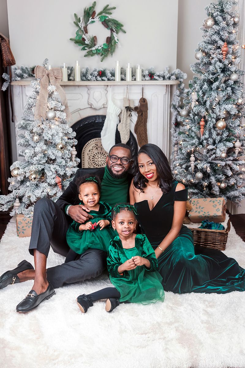 A joyful Black family in matching green velvet outfits poses together by a decorated Christmas fireplace and snow-flocked trees.